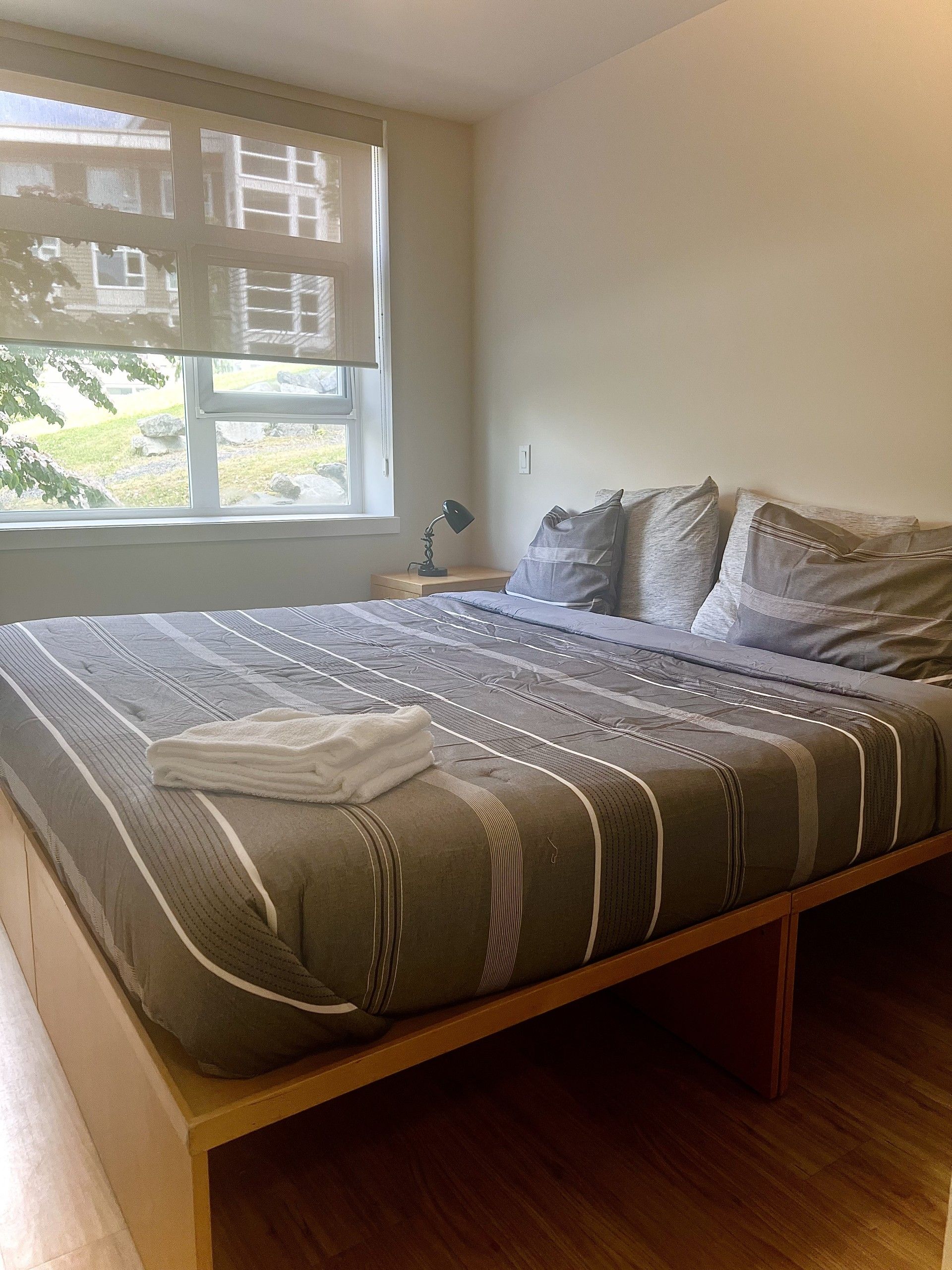 Bedroom with gray striped bedding, small side table, window with blind.