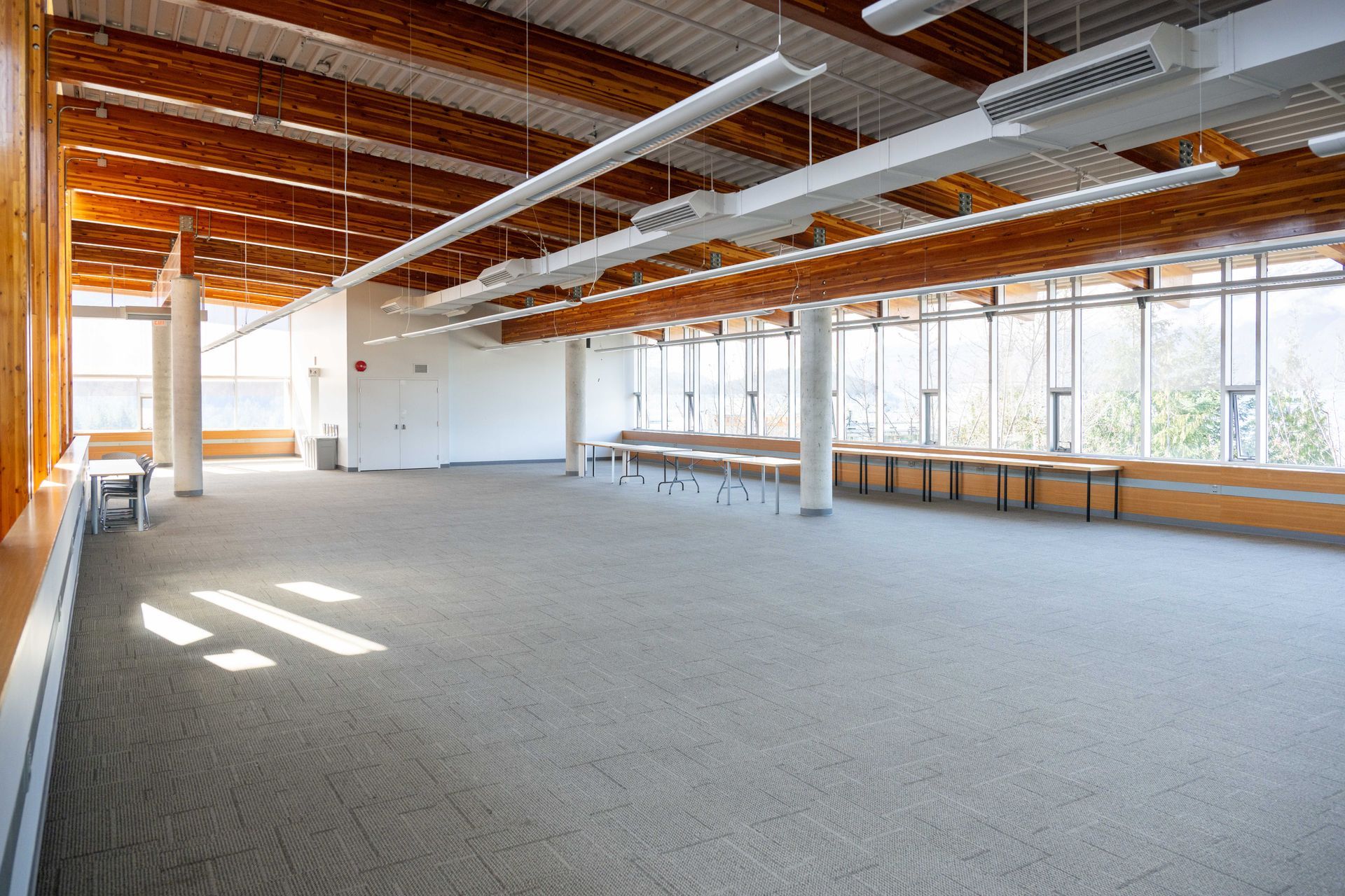 Empty modern office with carpeted floor, large windows, exposed ceiling beams, and desks along the wall