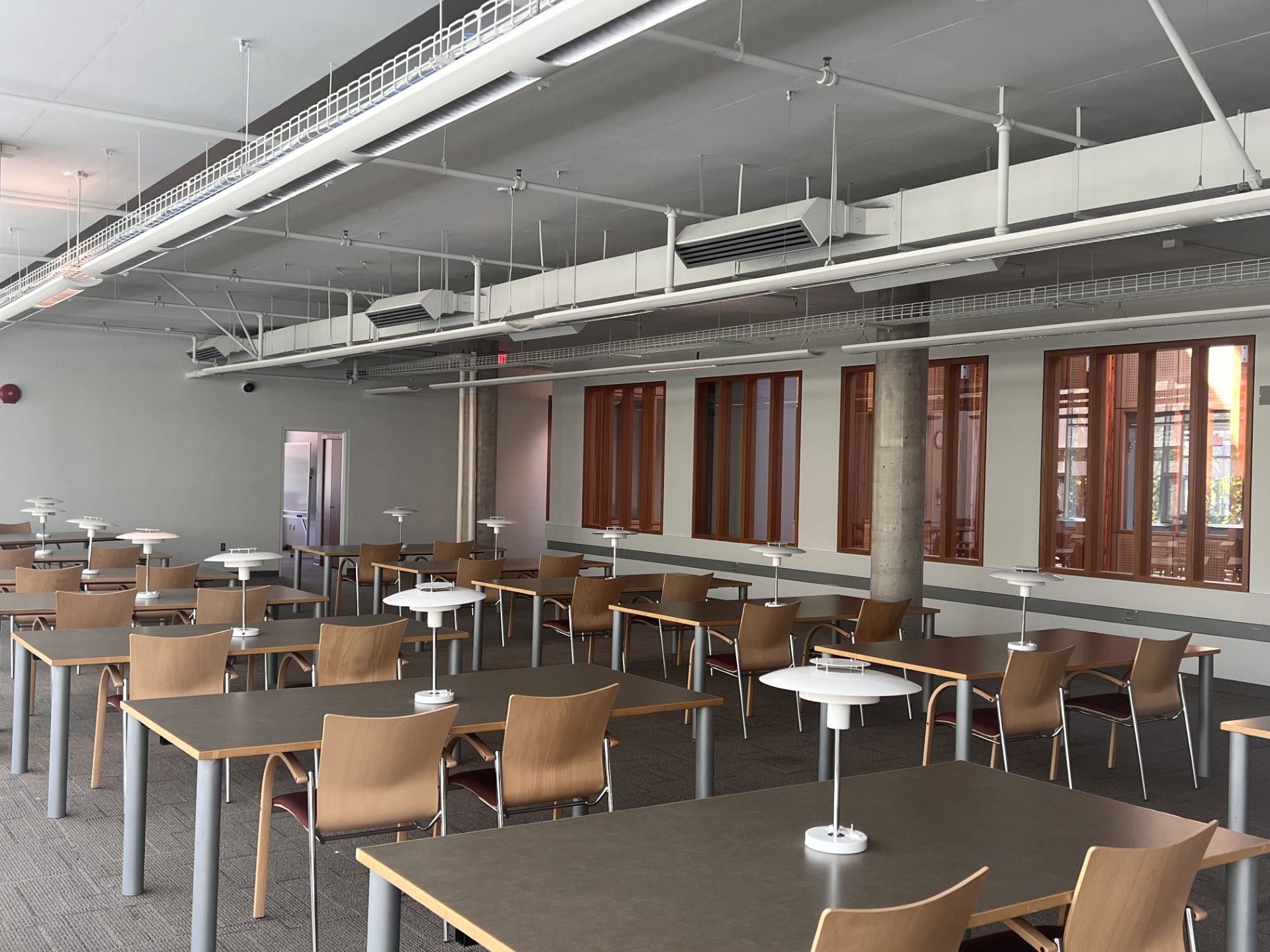 Empty modern classroom with rows of chairs and tables beneath exposed ceiling pipes and large windows