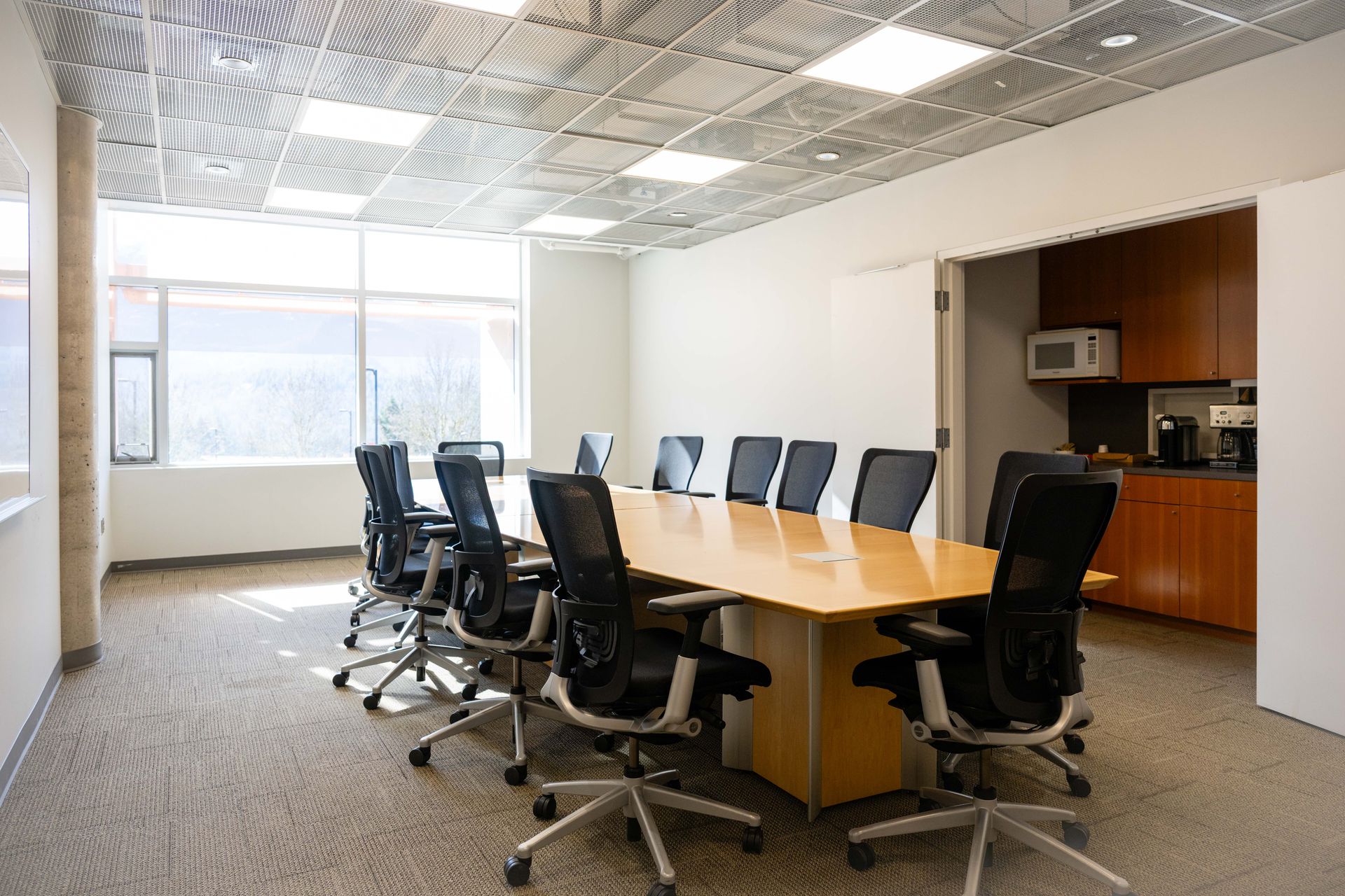 Empty conference room with long wood table, black office chairs, and bright windows.