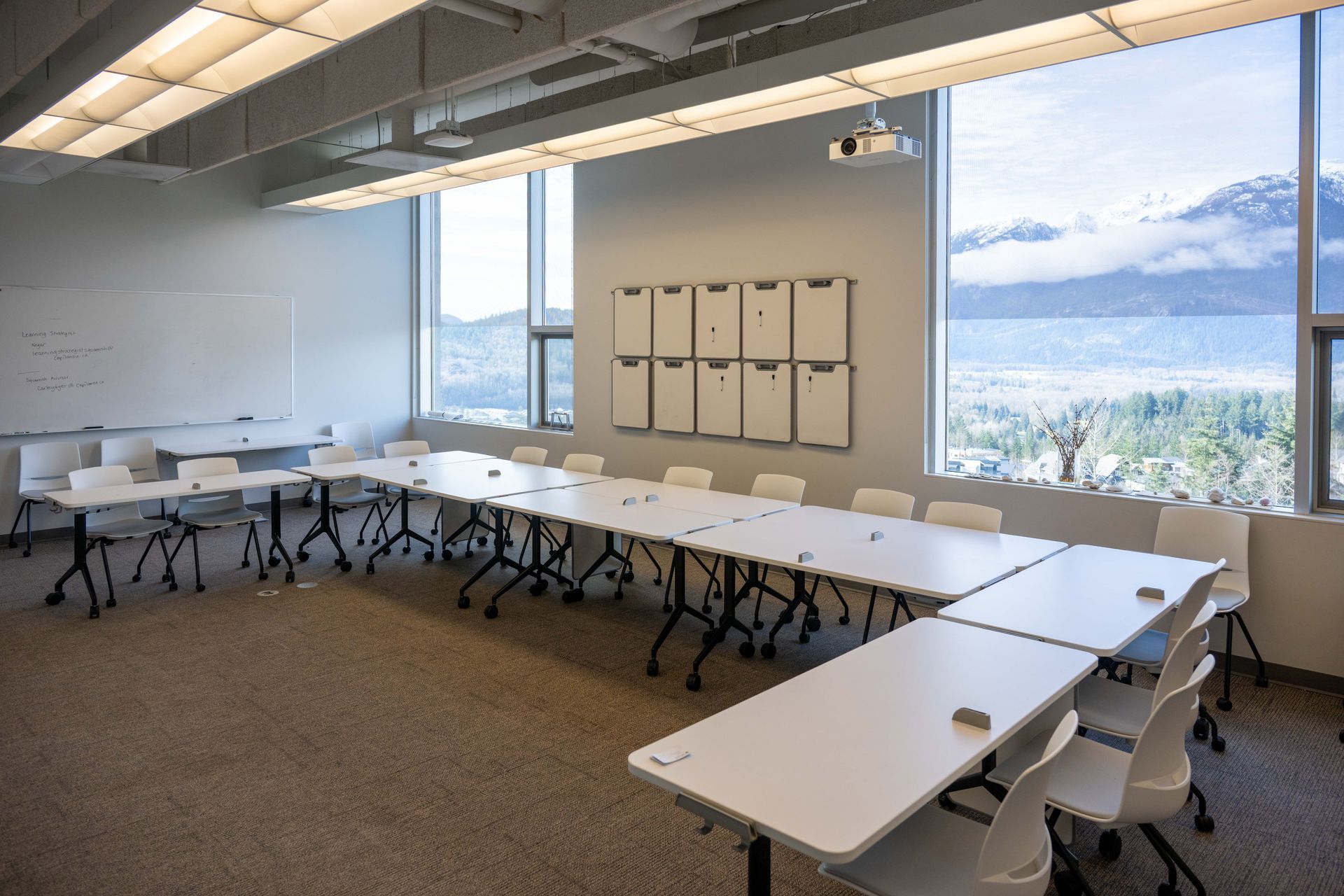 Modern meeting room with U-shaped white tables, chairs, windows, and wall-mounted whiteboards