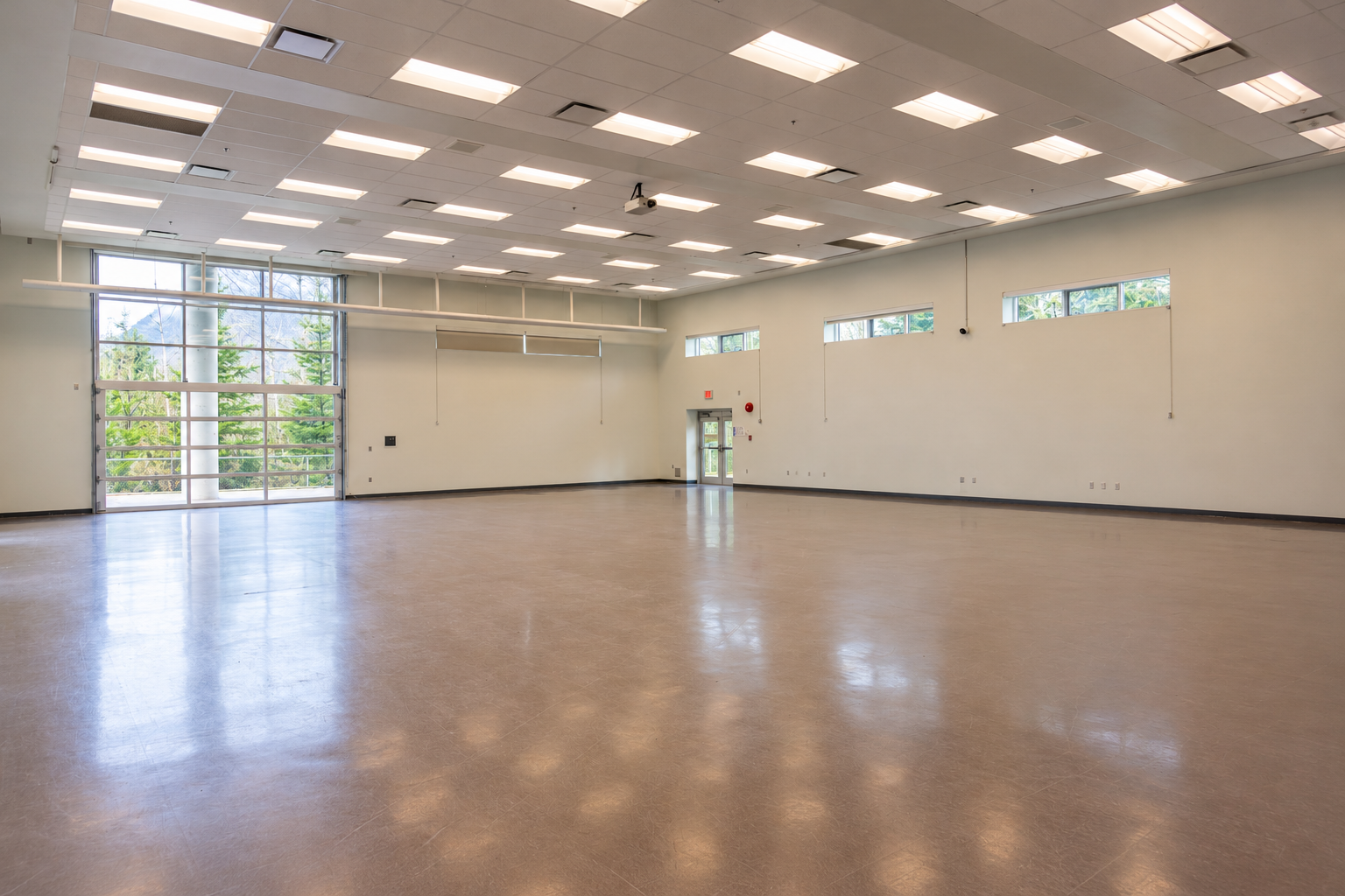 Empty bright community hall with polished floor, white walls, and large windows and ceiling lights