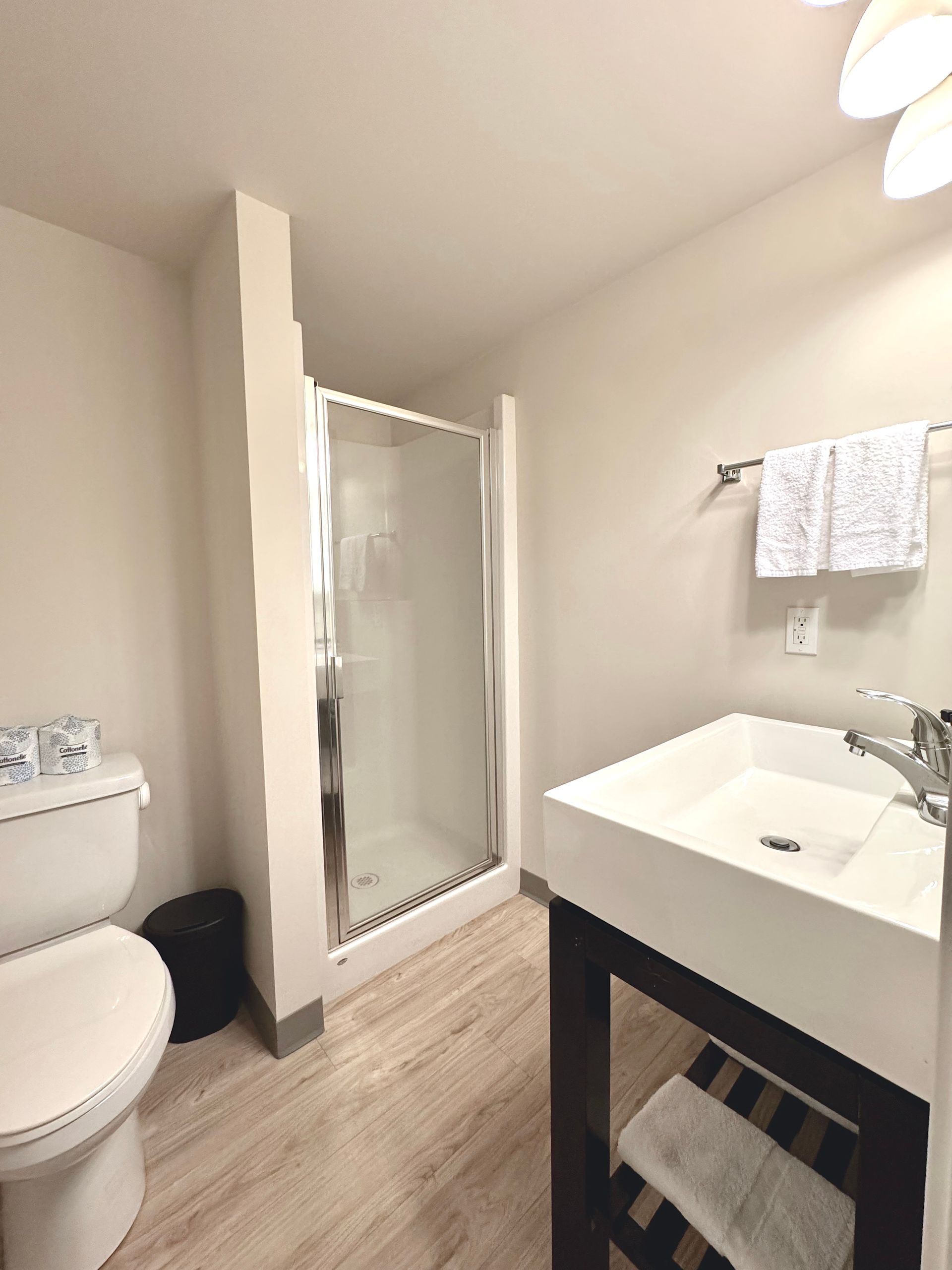 Small bathroom with toilet, sink, mirror, and towel rack on light walls and wood-look floor.
