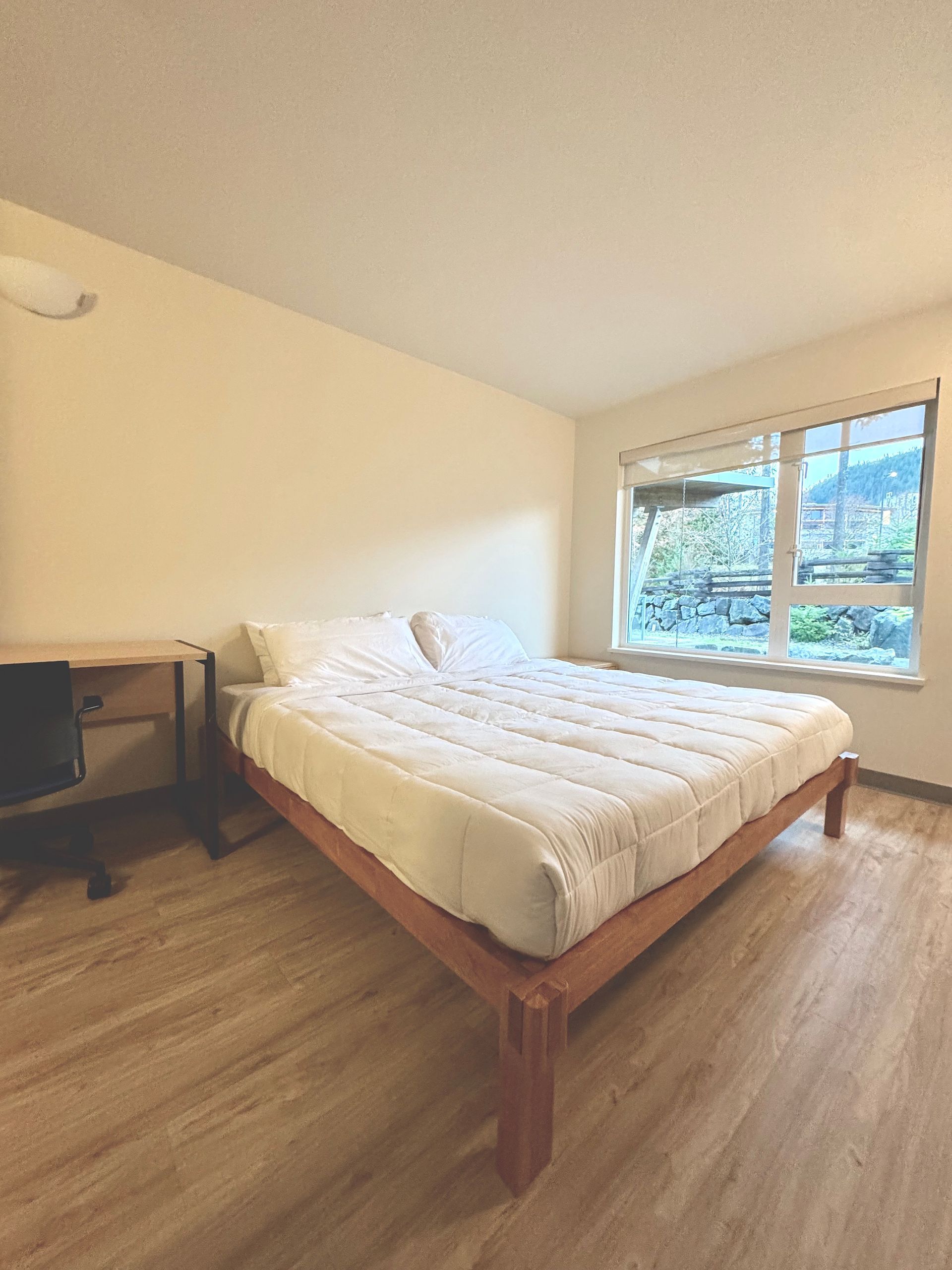 Bright bedroom with a bed, desk, and large window letting in daylight.