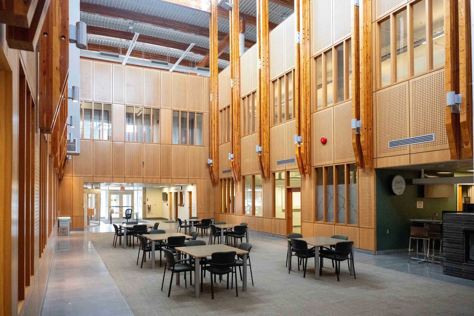 Modern wood-paneled atrium with tables and chairs under skylights, resembling a public lounge or cafeteria