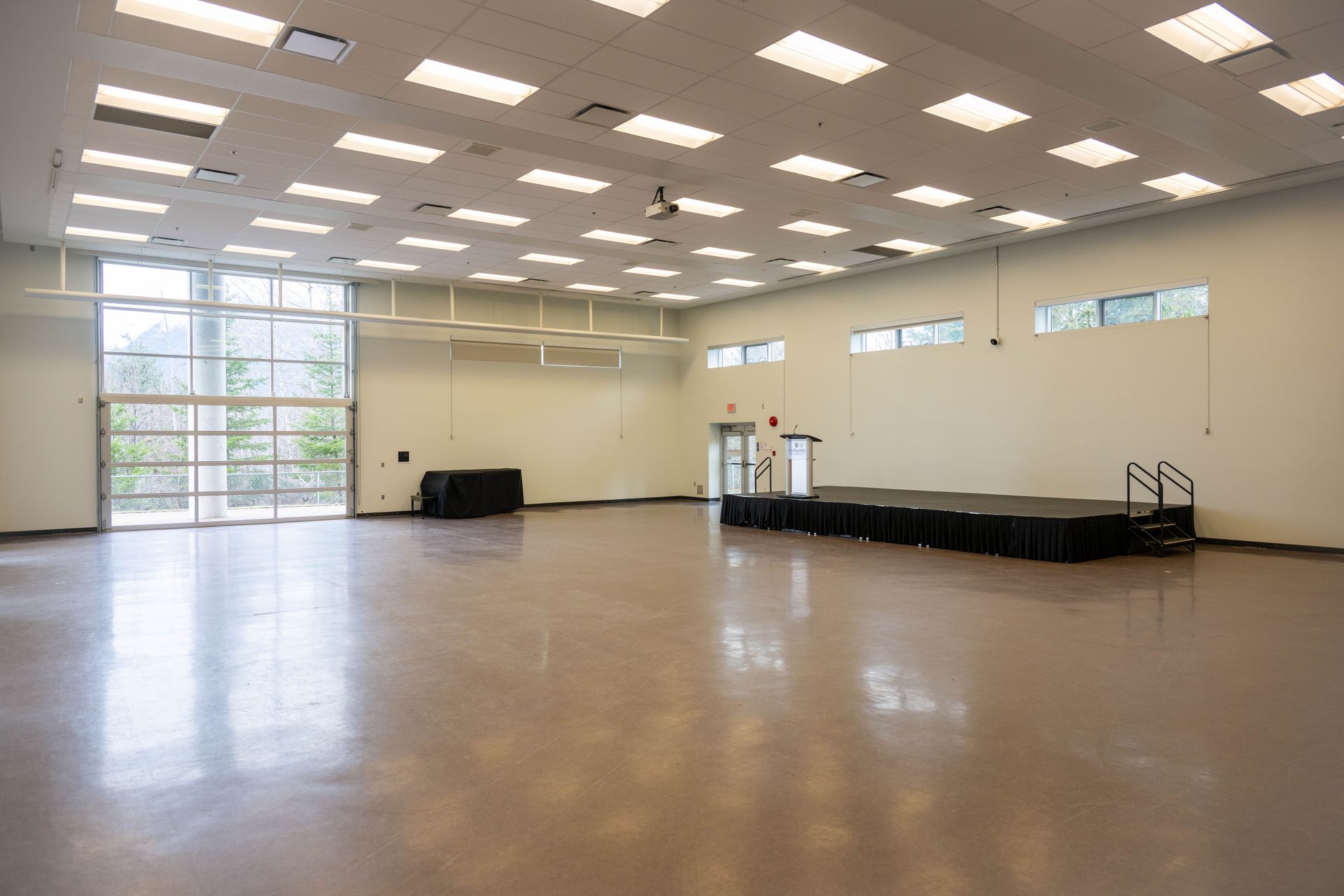 Empty indoor hall with polished floor, high ceiling lights, and a small raised stage at the far right.
