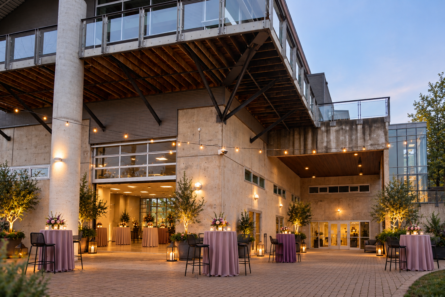 Outdoor evening venue with lit patio, tall columns, and cocktail tables under a modern building overhang