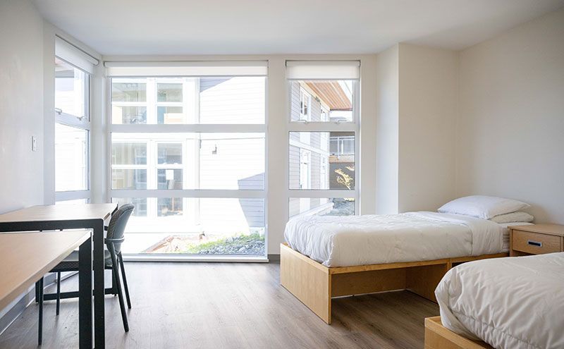 A bright dorm room with two beds, a desk, a chair, and a large window.