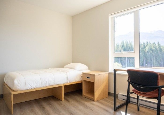 Empty, furnished dorm room with bed, nightstand, desk, chair, and window with mountain view.