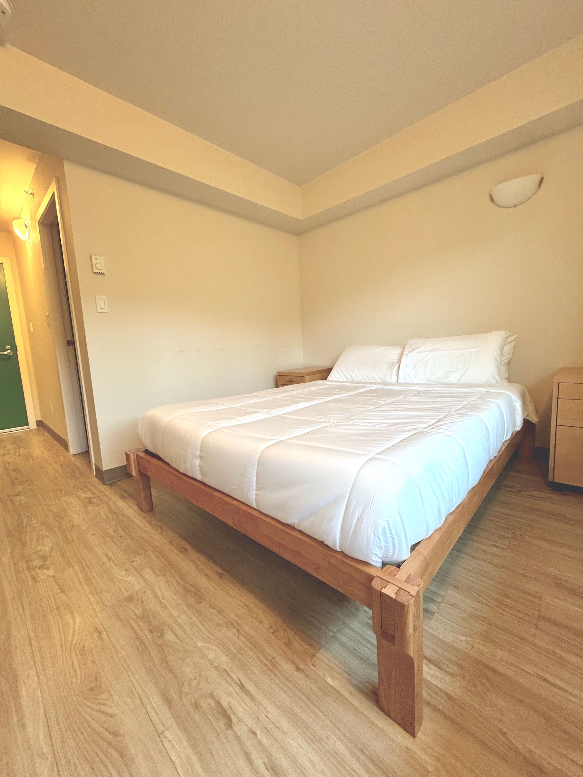 Bright bedroom with a wooden bed, white linens, and wood flooring under warm lighting