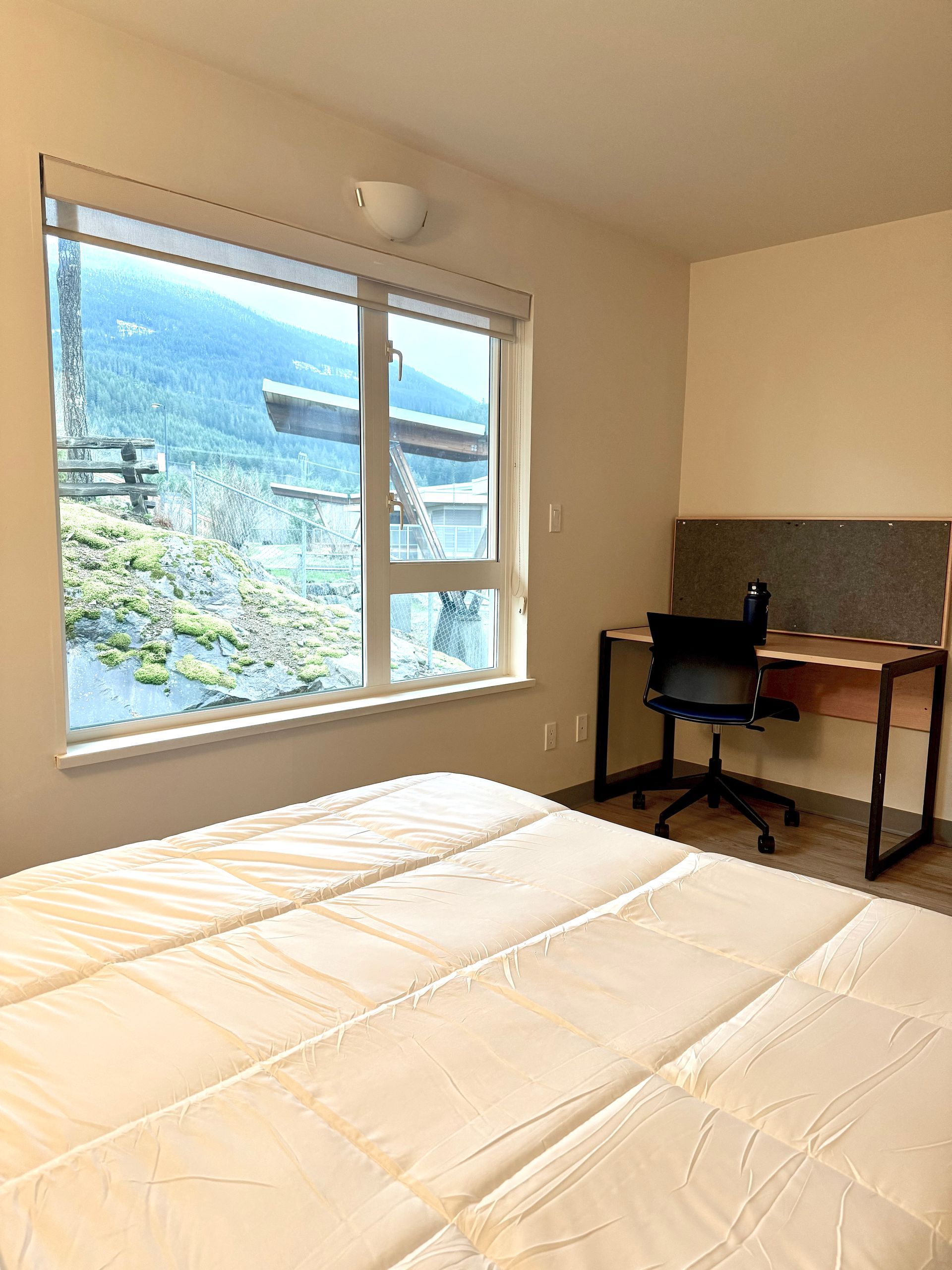 Bright bedroom with large window, bed, and desk overlooking a sunny green landscape