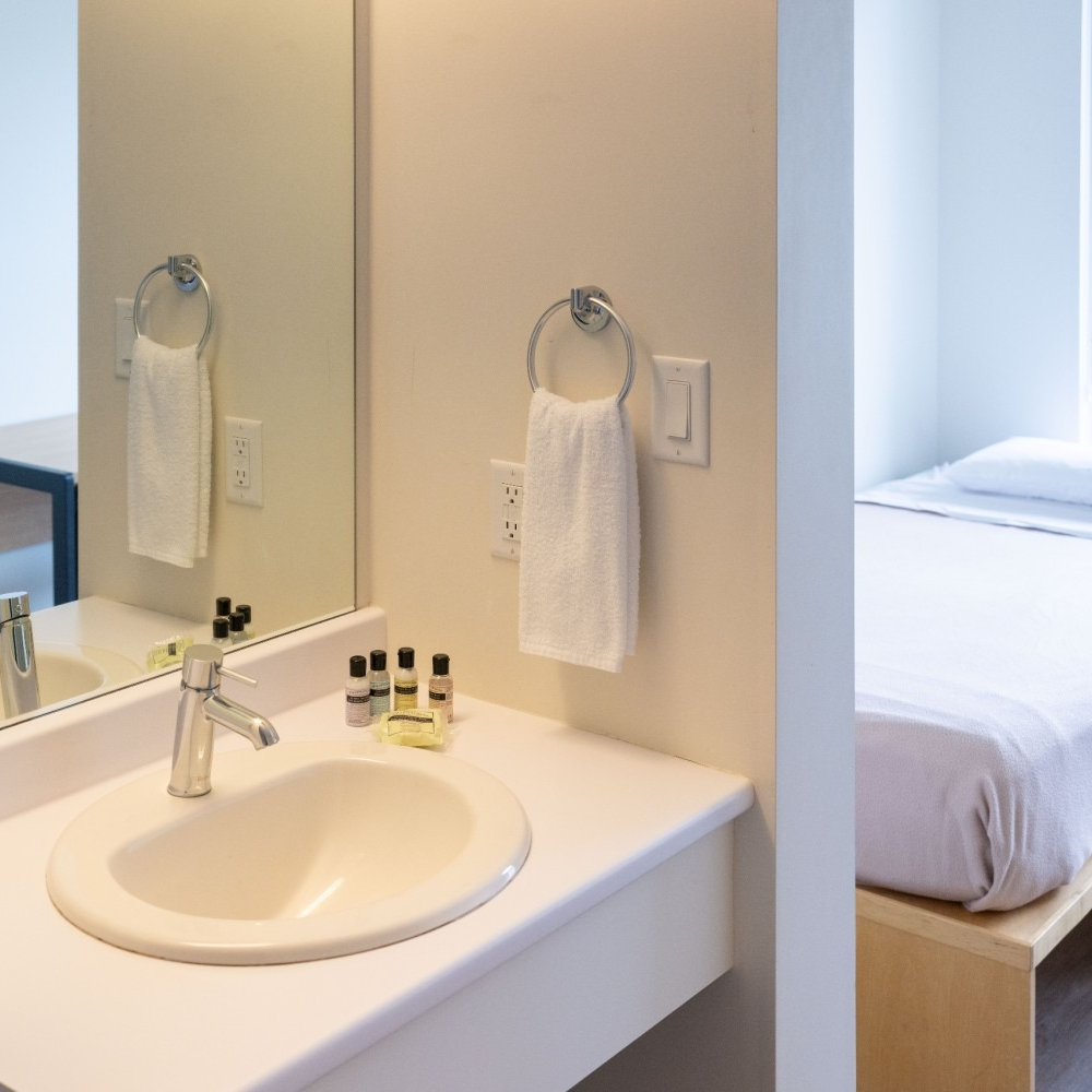 Bathroom with a sink, mirror, and towel rack. A bed is visible in the adjacent room.