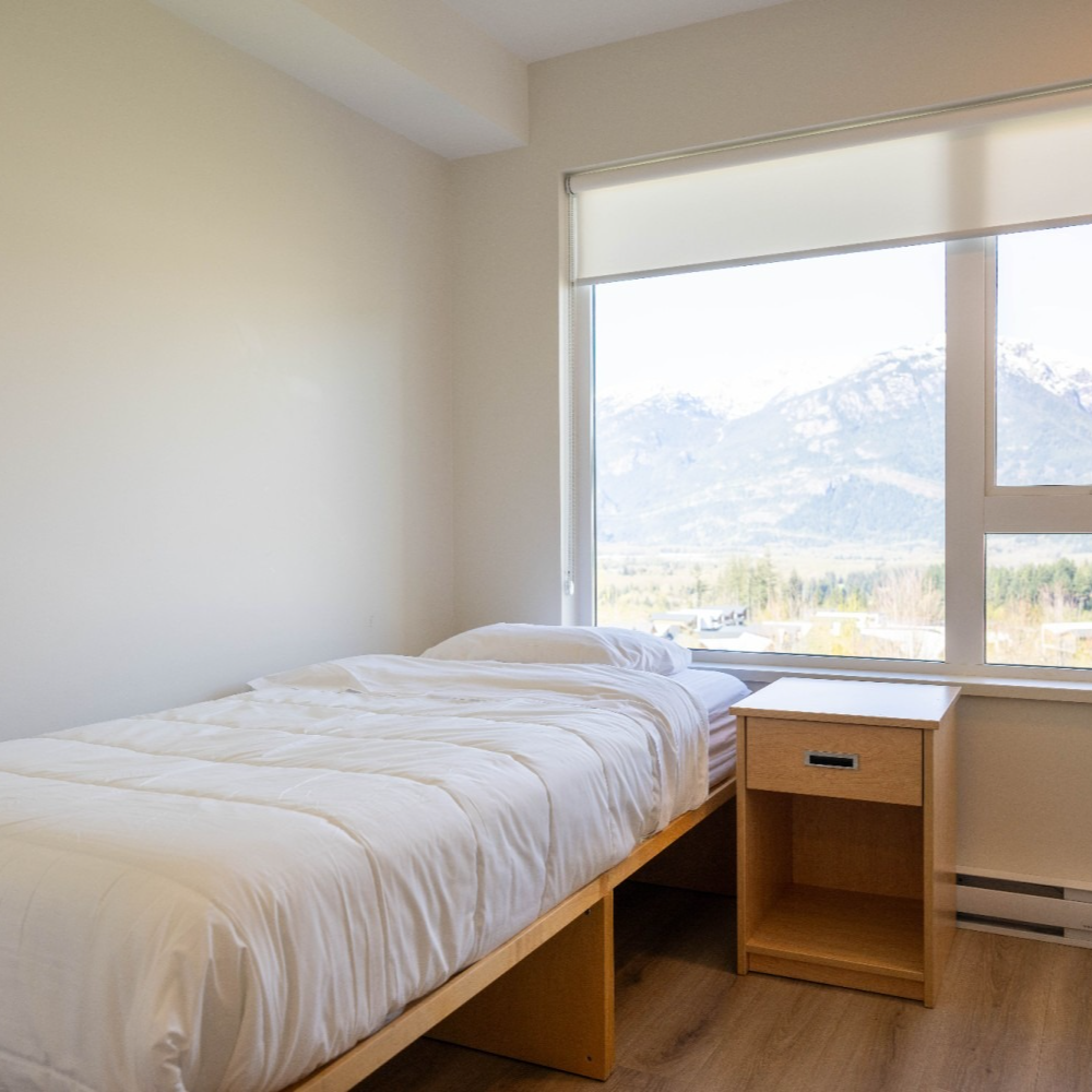 Bedroom with a single bed, nightstand, and window overlooking mountains. White walls and bedding.