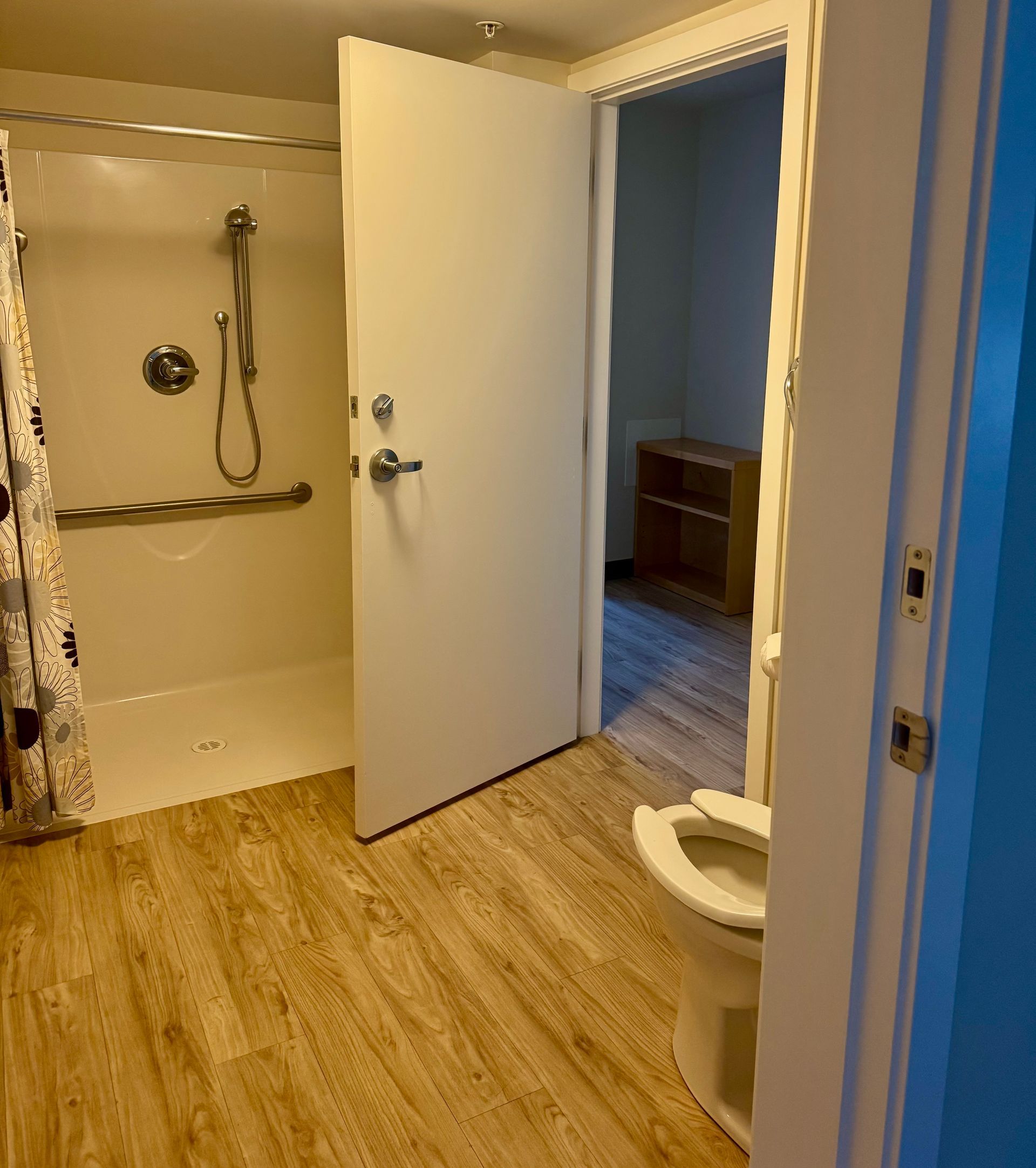 Open bathroom with shower, toilet, and wood-look flooring; door leads to a bedroom.