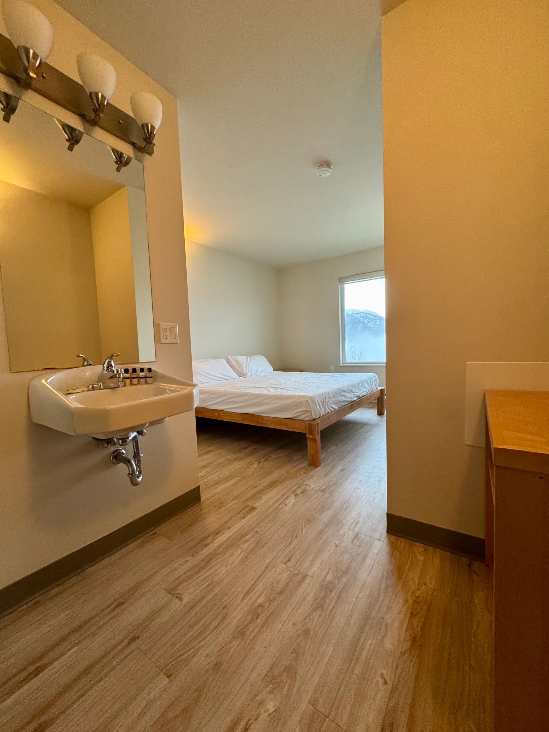 Small furnished bedroom with bed, sink, wooden floor, and window light