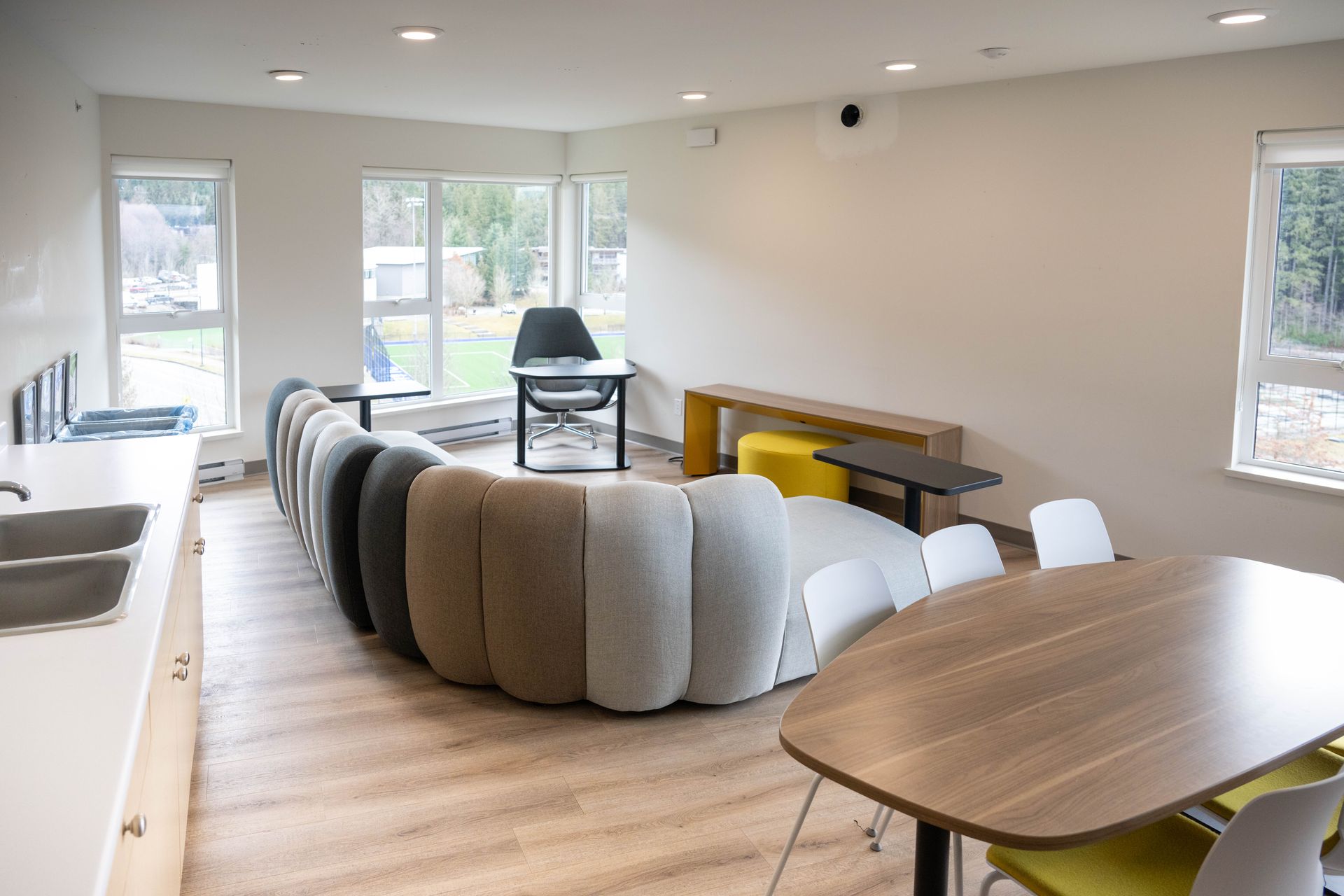 Spacious lounge with a curved sofa, small tables, and a dining table, with large windows overlooking greenery.