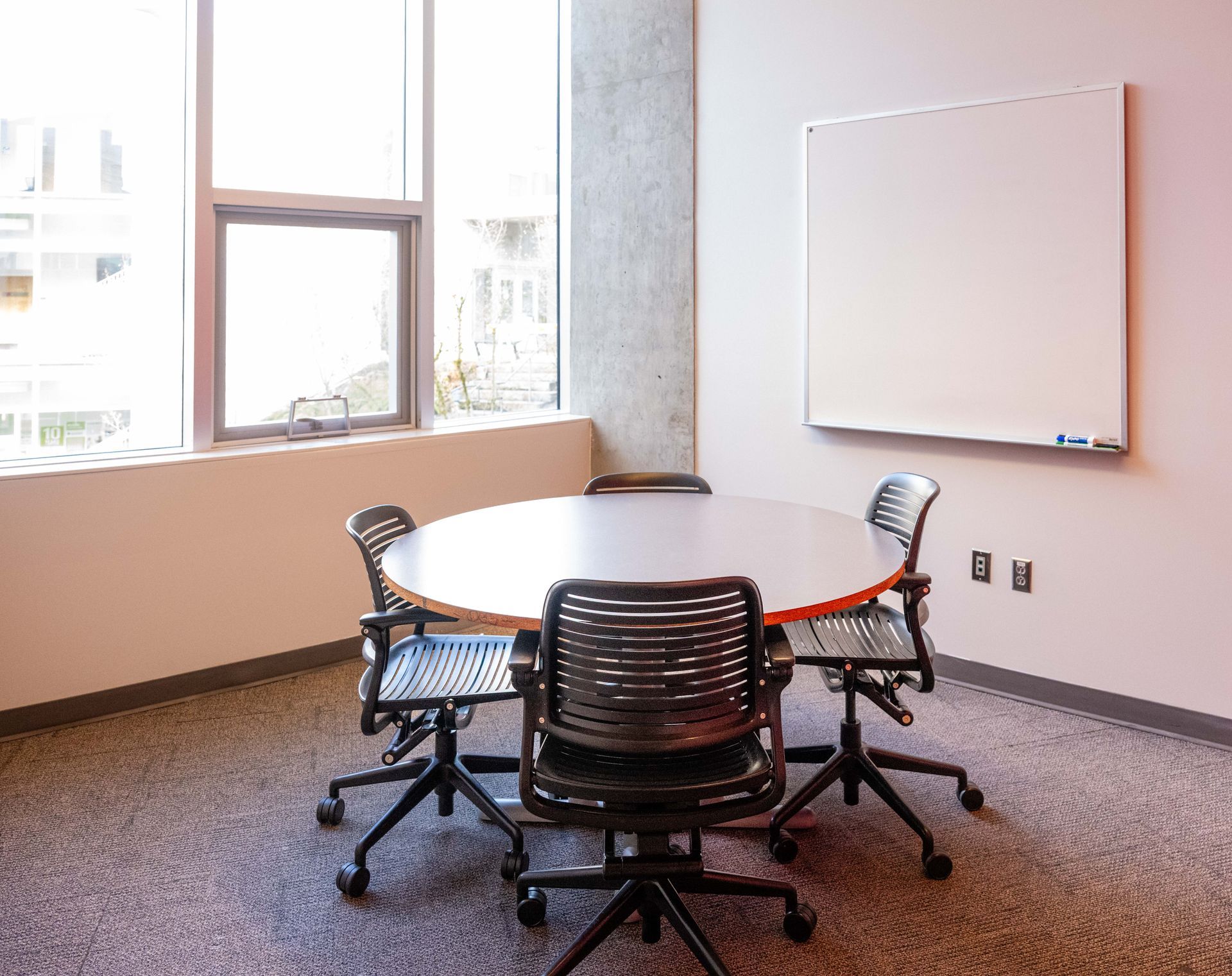 Small conference room with round table, four wheeled chairs, windows, and a whiteboard on the wall