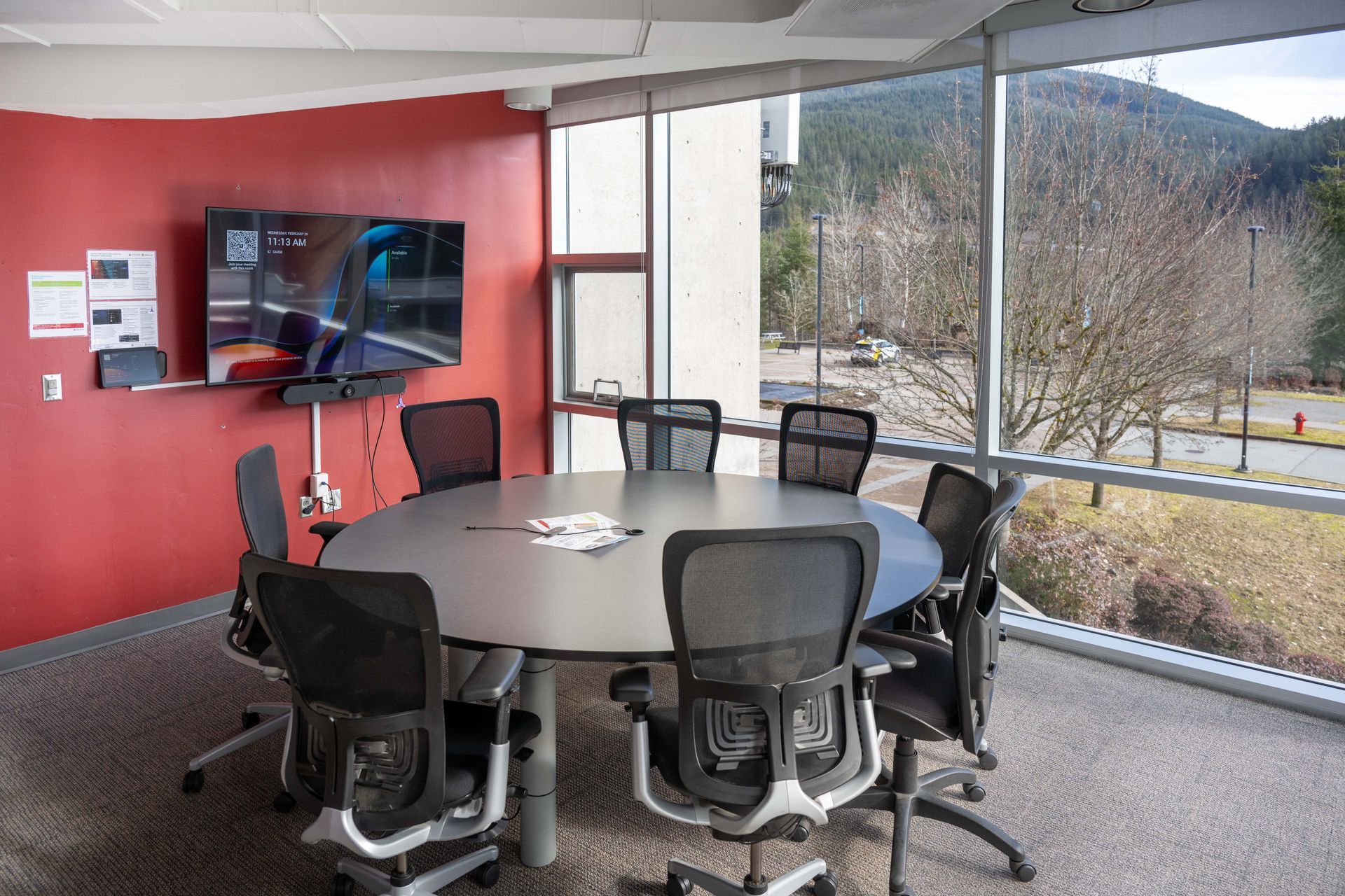 Conference room with oval table, office chairs, TV screen, and large windows overlooking trees and mountains
