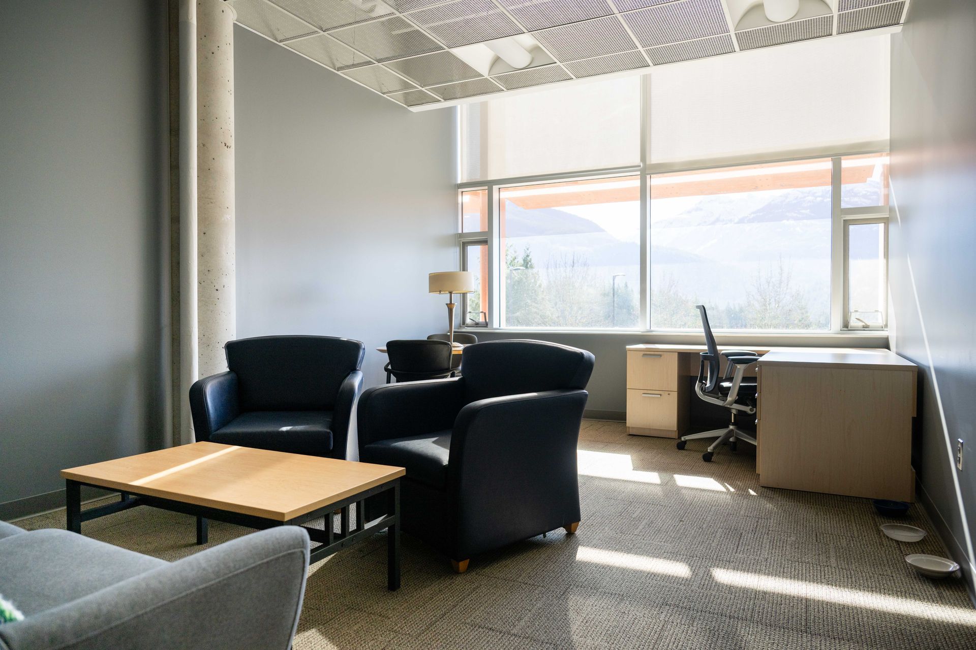 Empty office lounge with black chairs, wooden coffee table, and large windows letting in sunlight