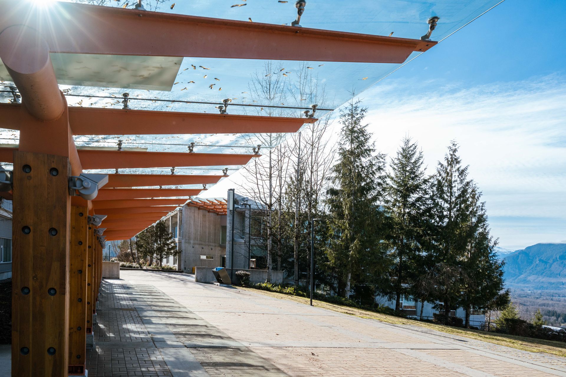 Pergola with glass roof, overlooking a pathway, trees, and distant mountain view on a sunny day.