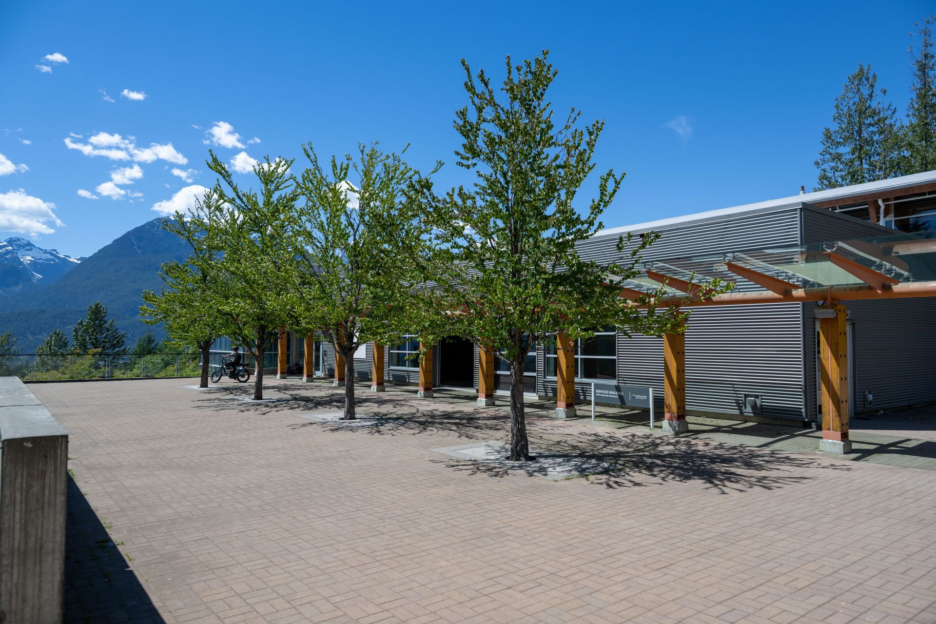 Modern outdoor plaza with young trees, mountain backdrop, and a covered walkway under a blue sky