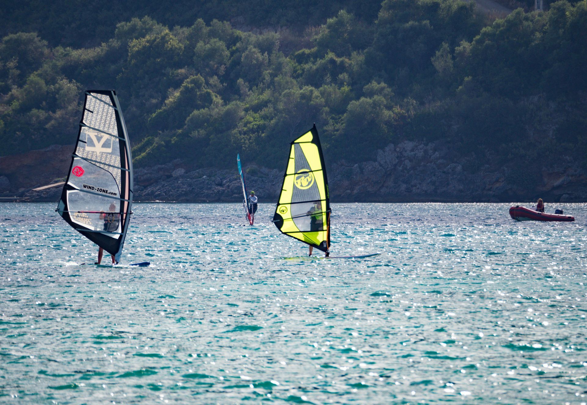 Windsurfers on the water with sails up; small boat in background, trees on hillside.