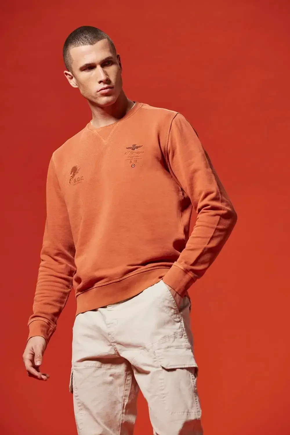 En mand iført sweater og khakibukser står foran en rød baggrund.