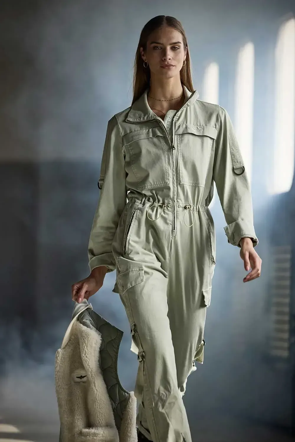 En kvinde i en jumpsuit står i et rum med en jakke i hånden.