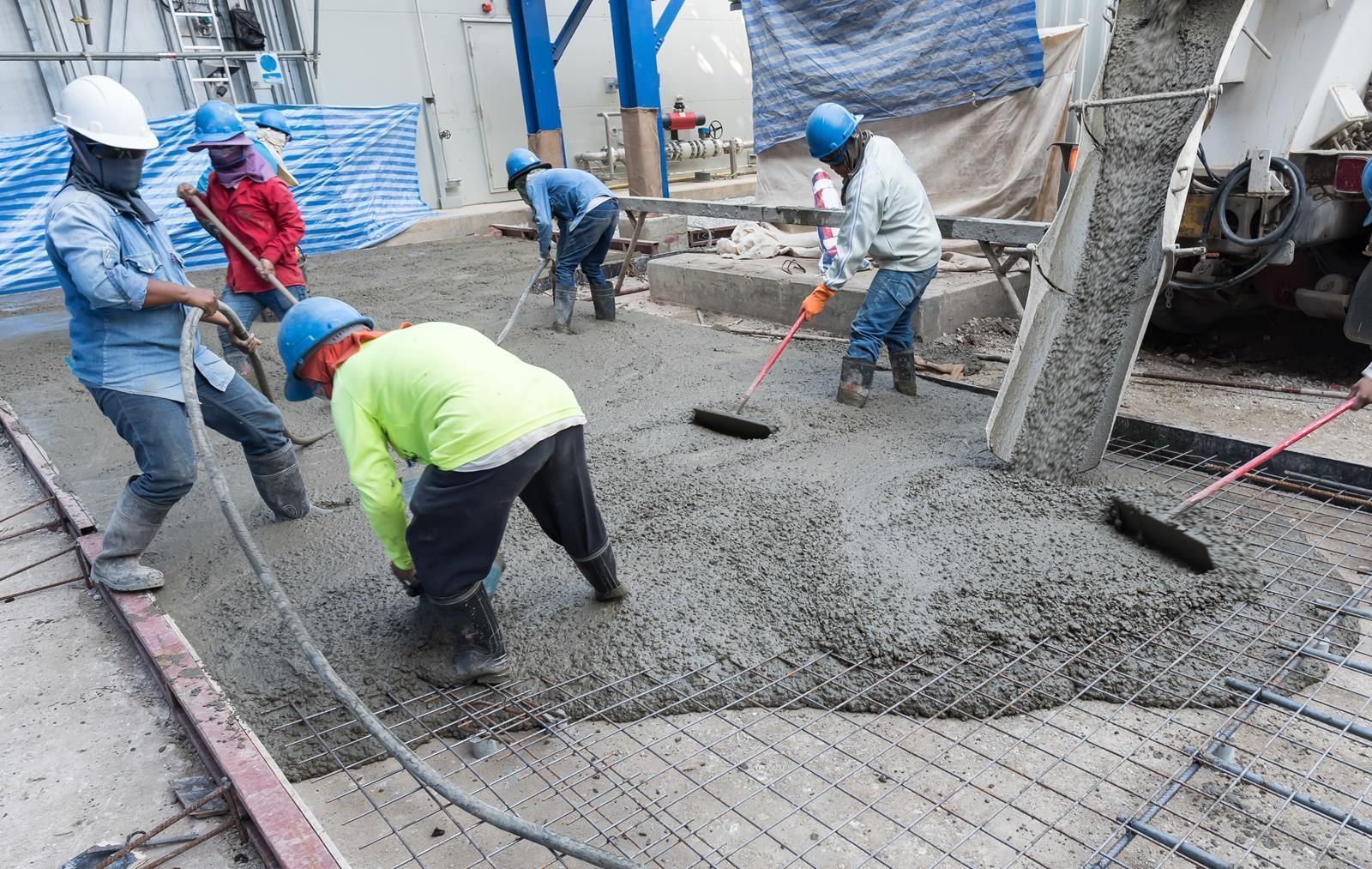 Construction workers smoothing wet concrete on a site with a mixer truck and blue-covered materials