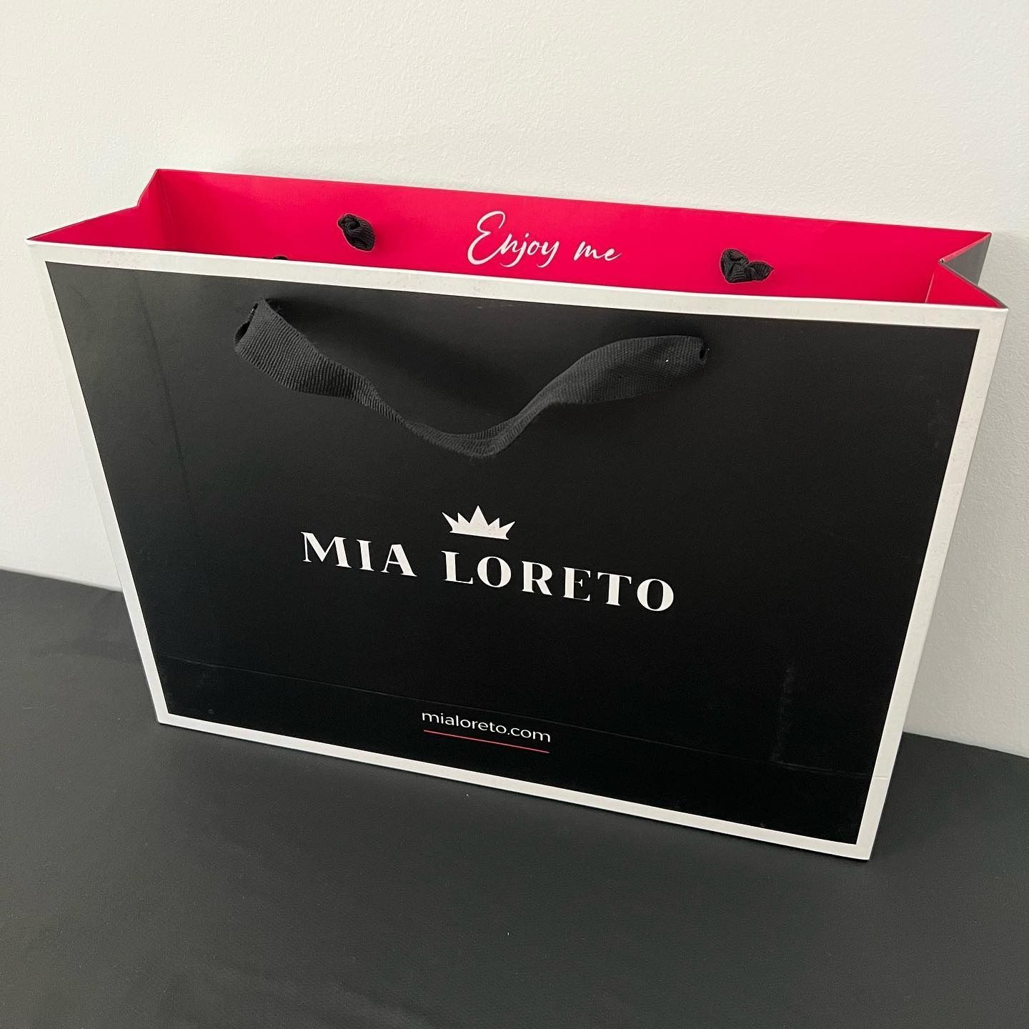 Bolsa de regalo negra de Mia Loreto con parte superior rosa y asas de cinta negra sobre una superficie gris.