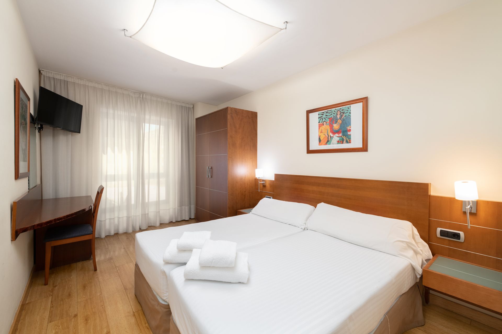Habitación de hotel luminosa con cama doble, muebles de madera, escritorio, televisor de pared y cortinas translúcidas.