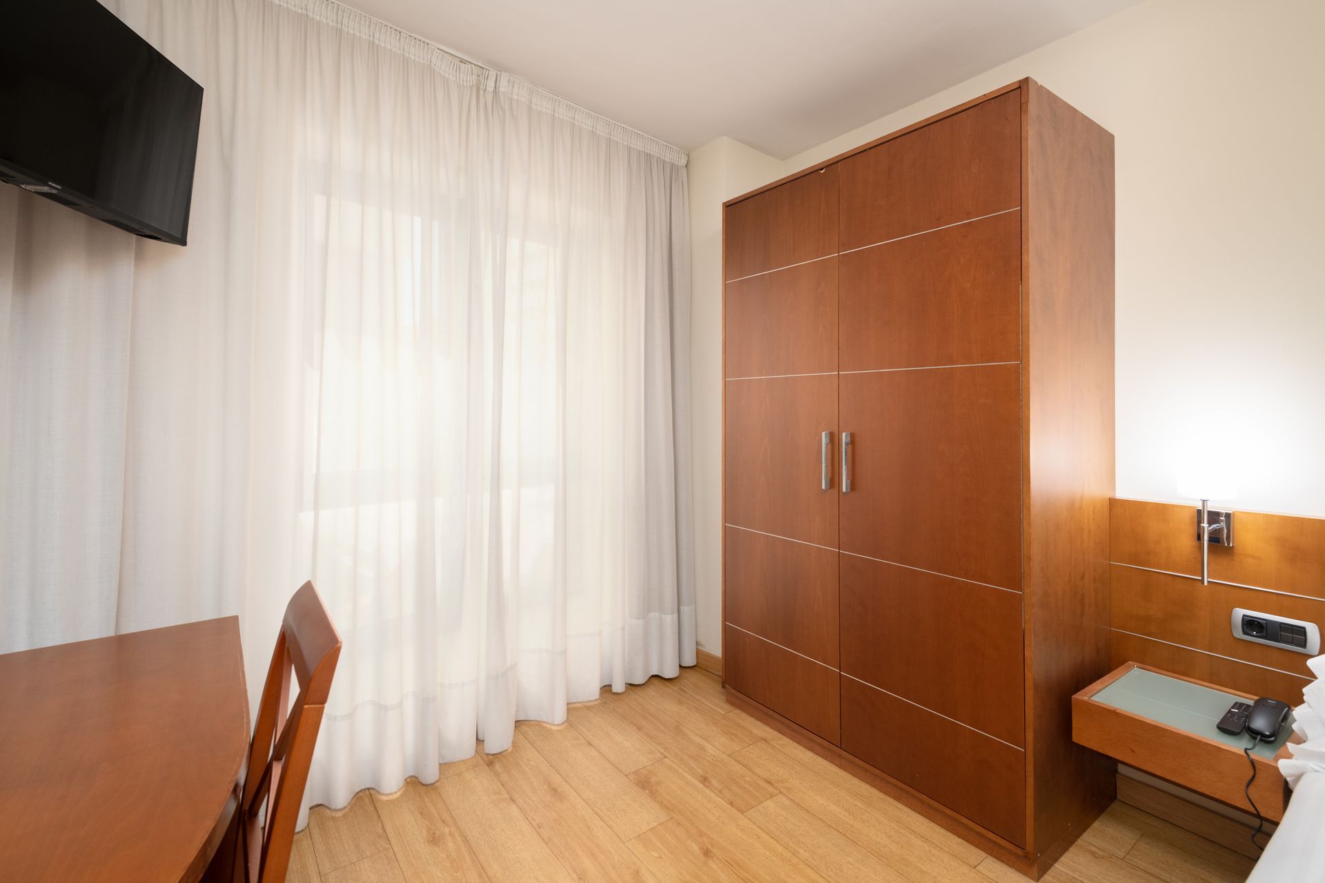 Dormitorio pequeño con armario de madera, escritorio, televisor montado en la pared y cortinas transparentes en una ventana.