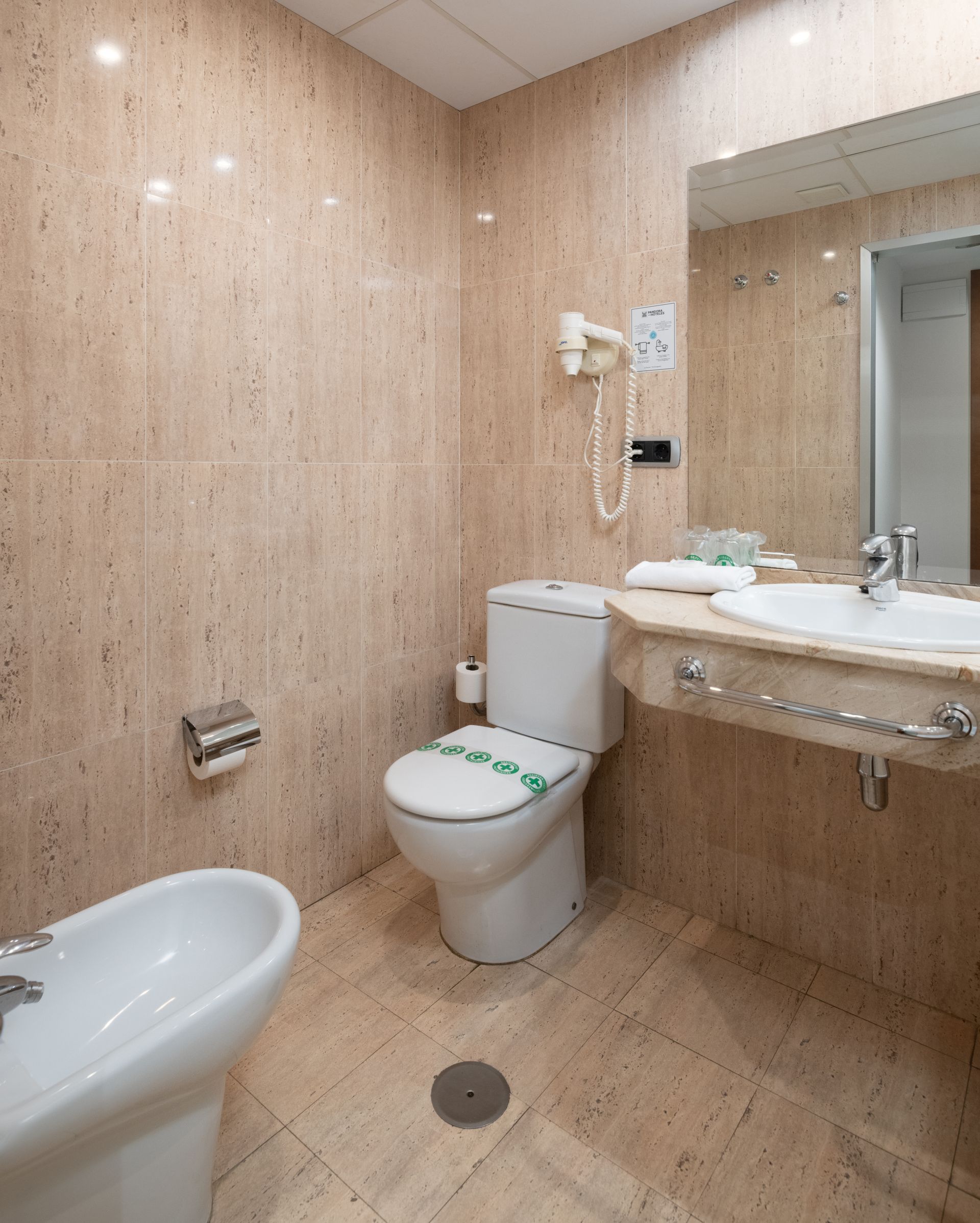 Pequeño baño alicatado en tonos beige, con inodoro, lavabo, bidé, espejo y manguera de ducha
