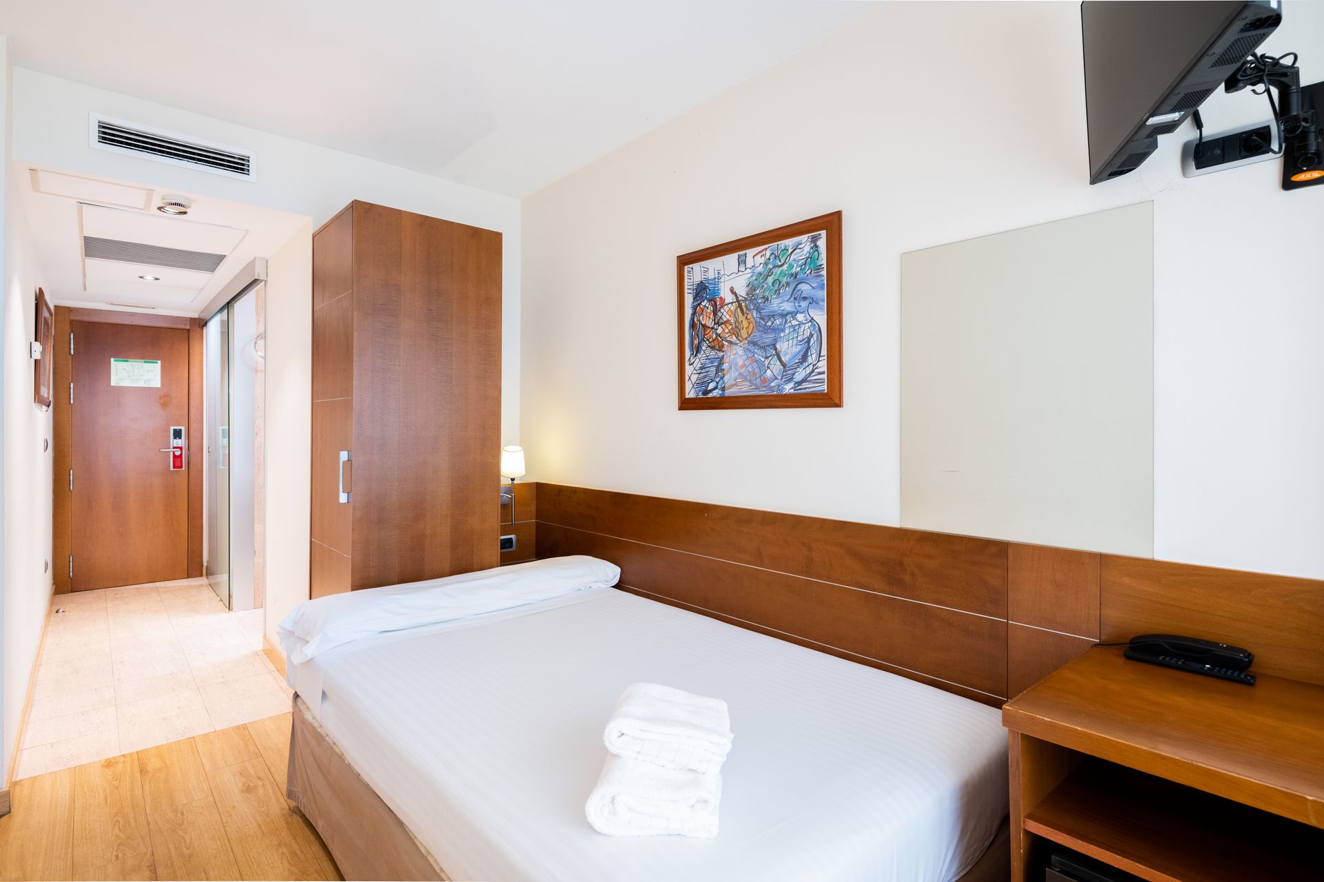Habitación pequeña de hotel con dos camas individuales, muebles de madera, cuadros en las paredes y vistas a un pasillo.