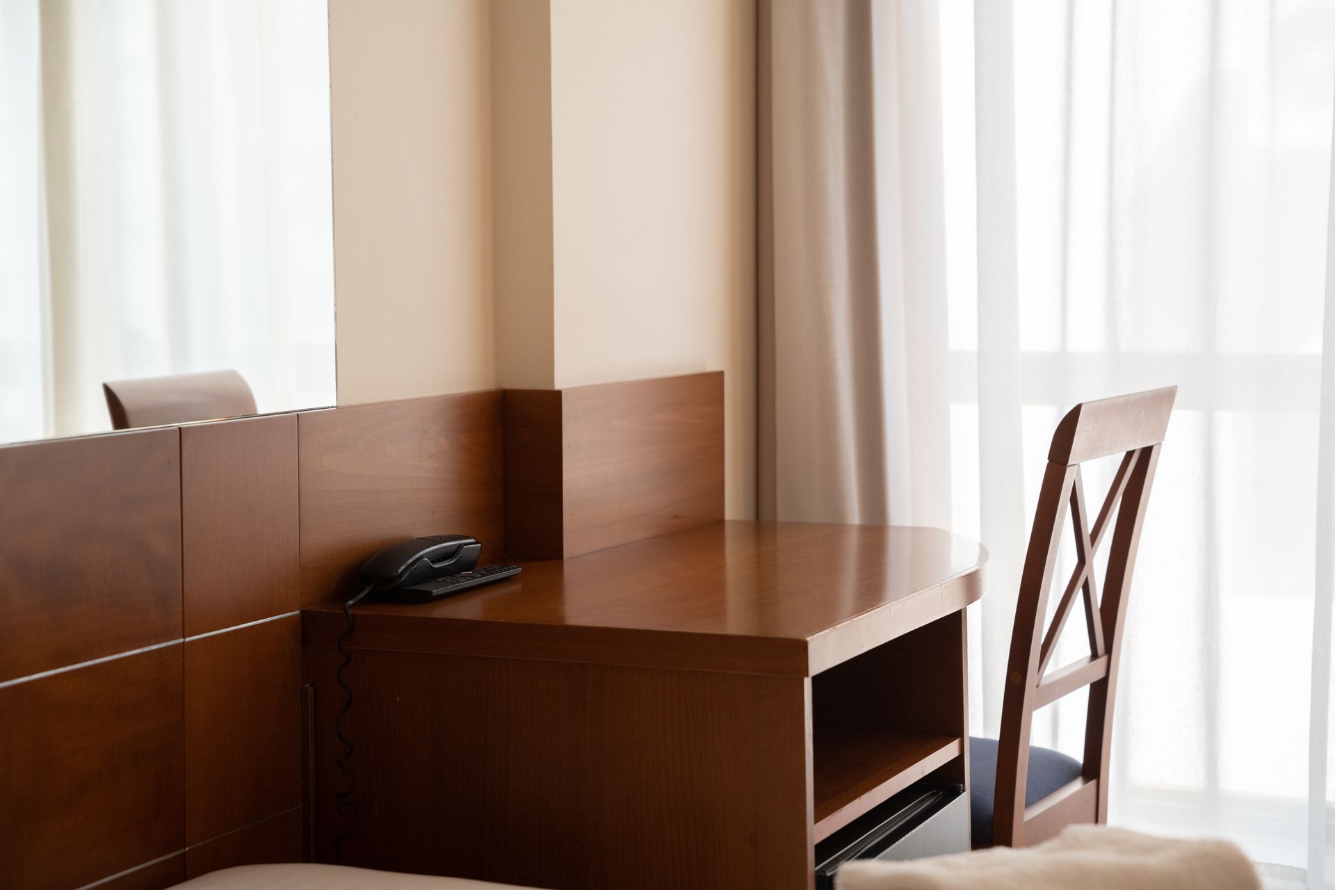 Escritorio de madera con un teléfono negro junto a una silla, en una habitación luminosa con cortinas translúcidas