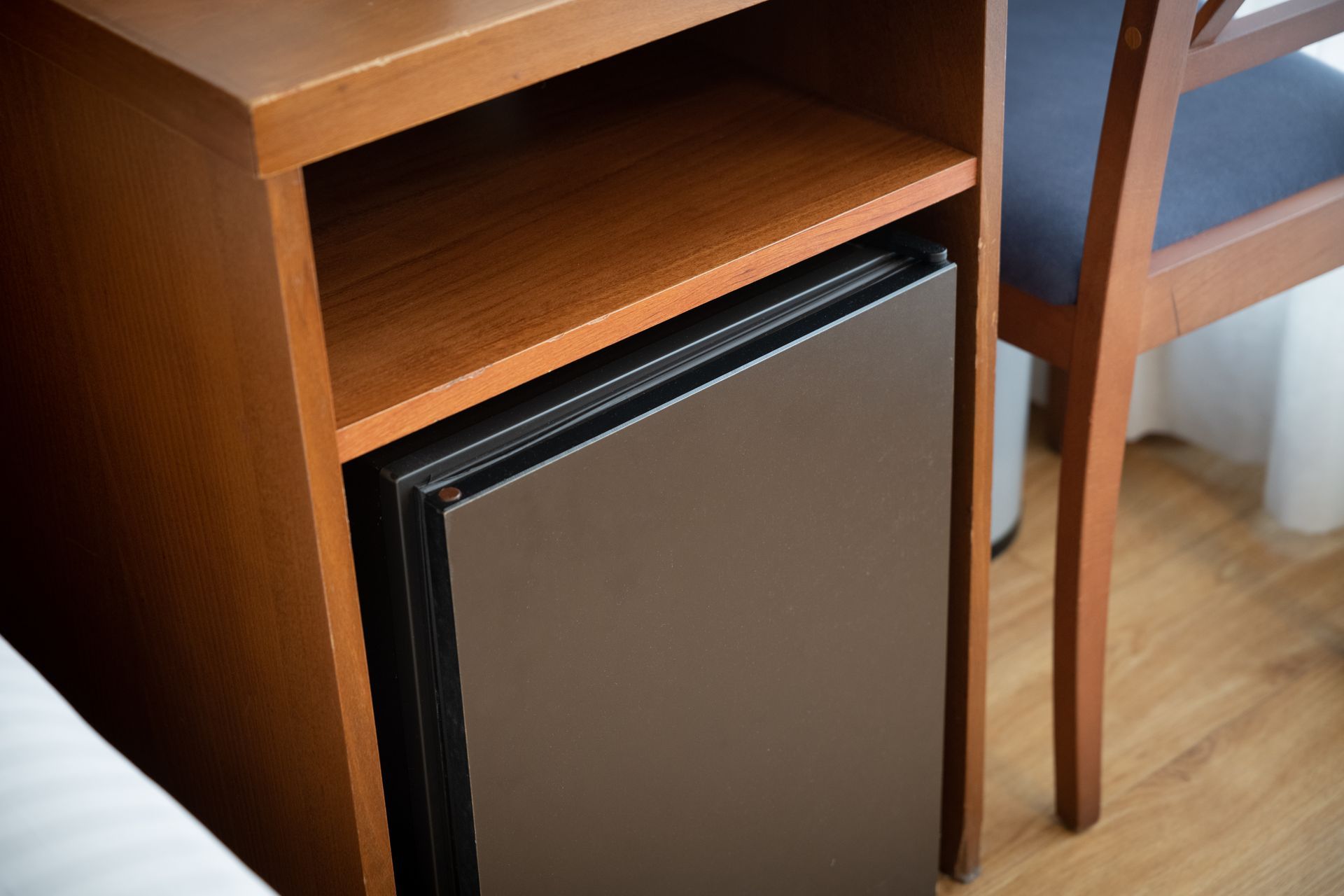 Mini nevera negra pequeña debajo de una mesa auxiliar de madera, junto a una silla, sobre un suelo de madera dura