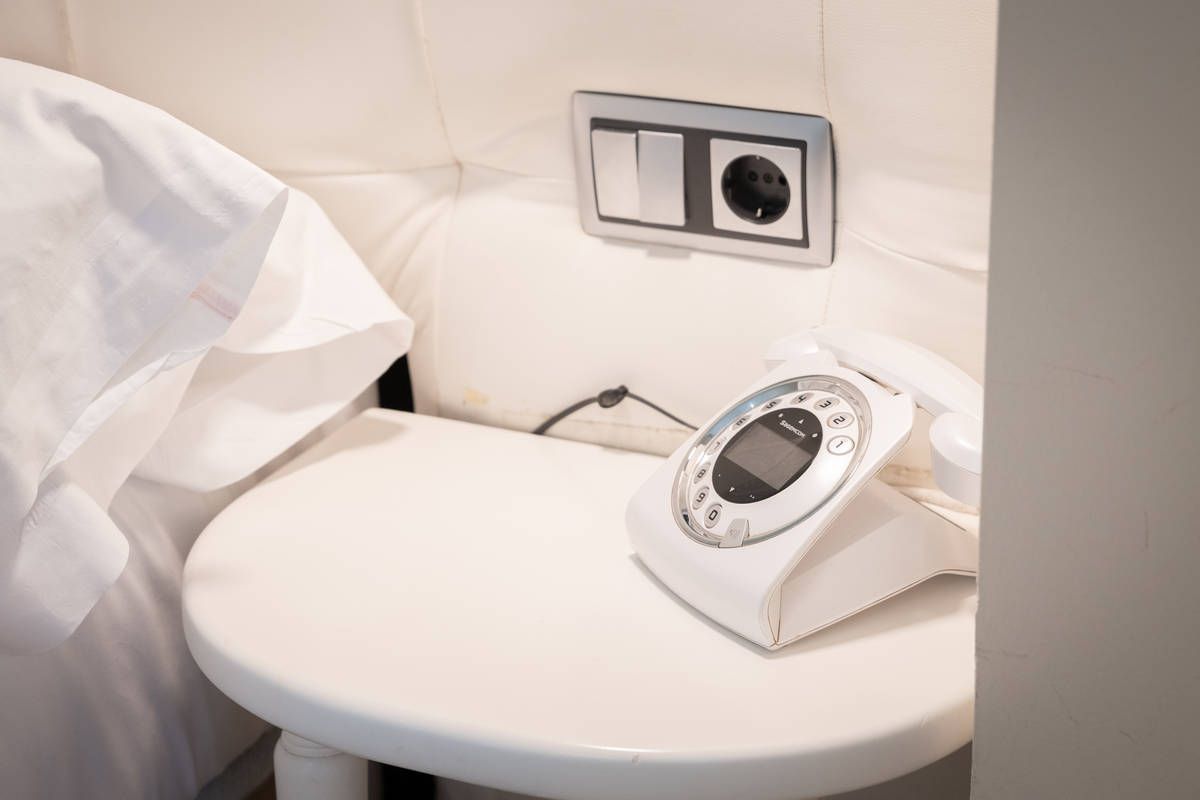 Mesita de noche pequeña y blanca con un teléfono con cable y un enchufe de pared en una habitación de aspecto hotelero