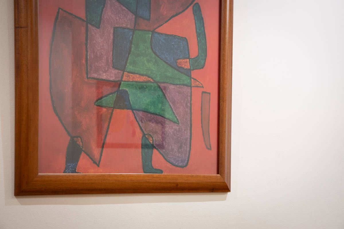 Cuadro abstracto enmarcado en madera, con formas geométricas rojas, verdes y moradas, colgado en una pared color crema