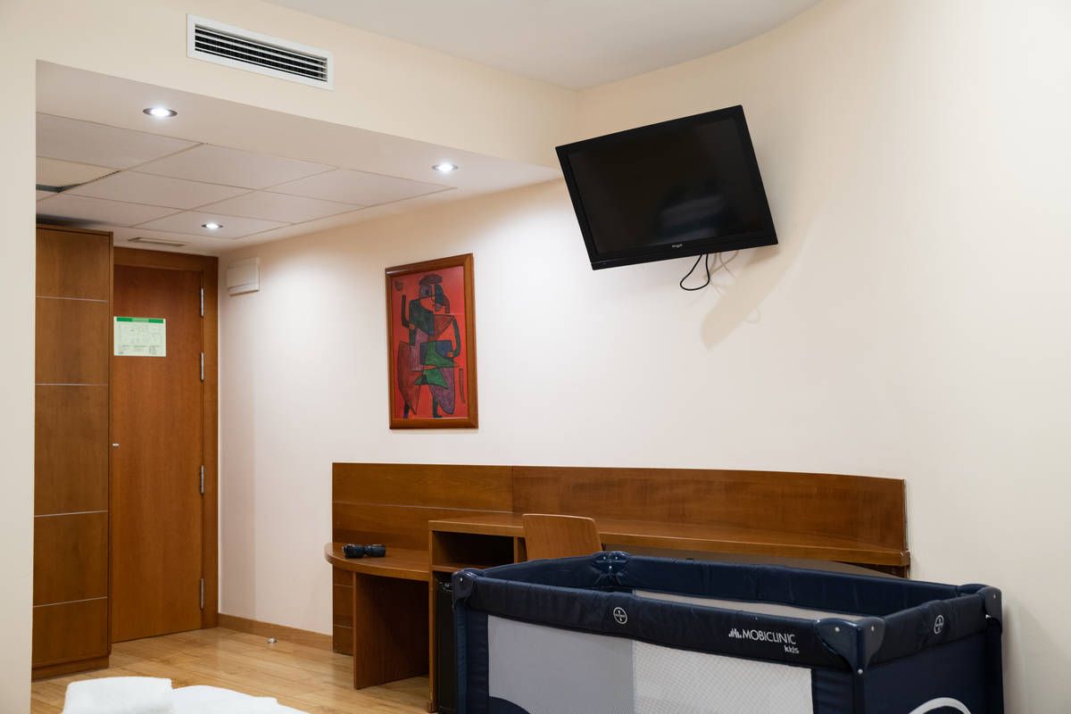 Habitación de hospital con televisor instalado en la pared, banco de madera y cortina de privacidad alrededor de una cama