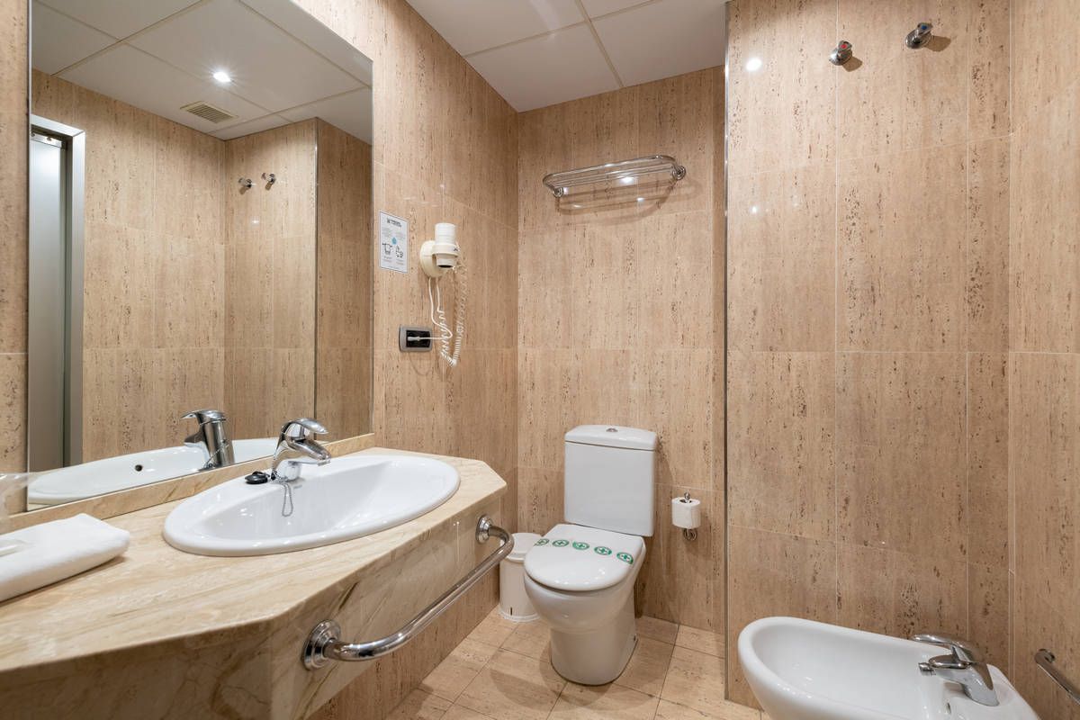 Baño moderno con azulejos beige, lavabo, inodoro, espejo y bidé.