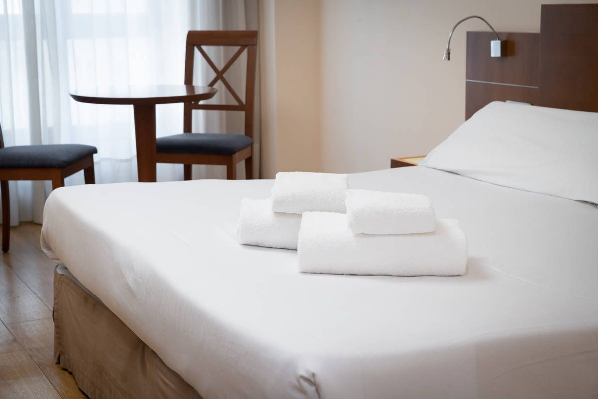 Cama de hotel impecablemente hecha, con toallas blancas dobladas en una habitación luminosa con sillas junto a la ventana
