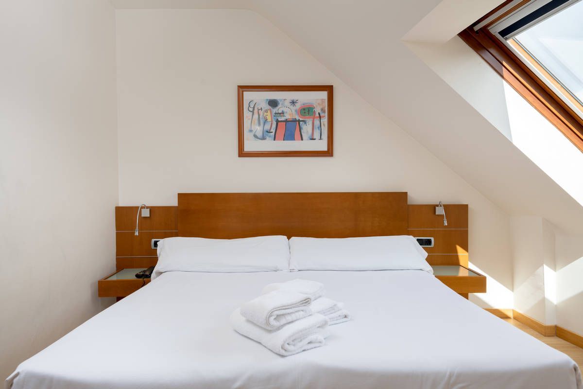 Dormitorio luminoso con una cama blanca, un cabecero de madera y ventanas de techo inclinadas.