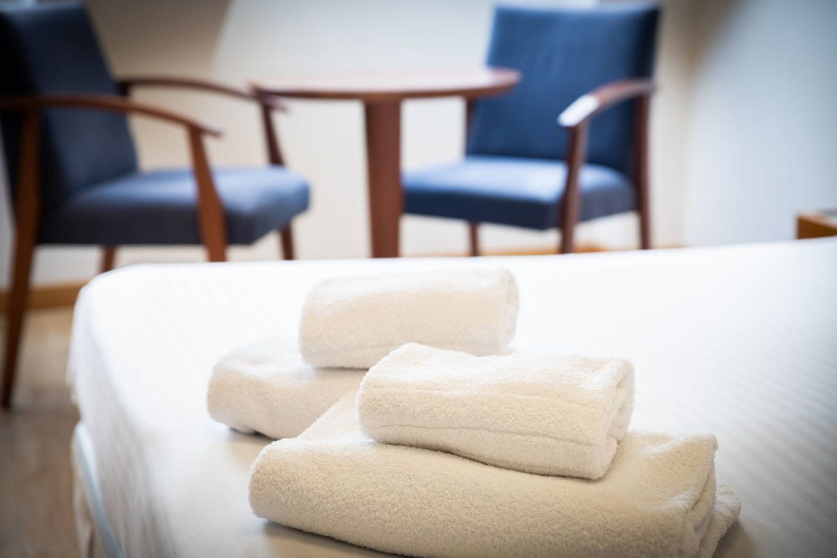 Toallas blancas dobladas sobre una cama en una habitación de hotel, con dos sillas y una mesita al fondo