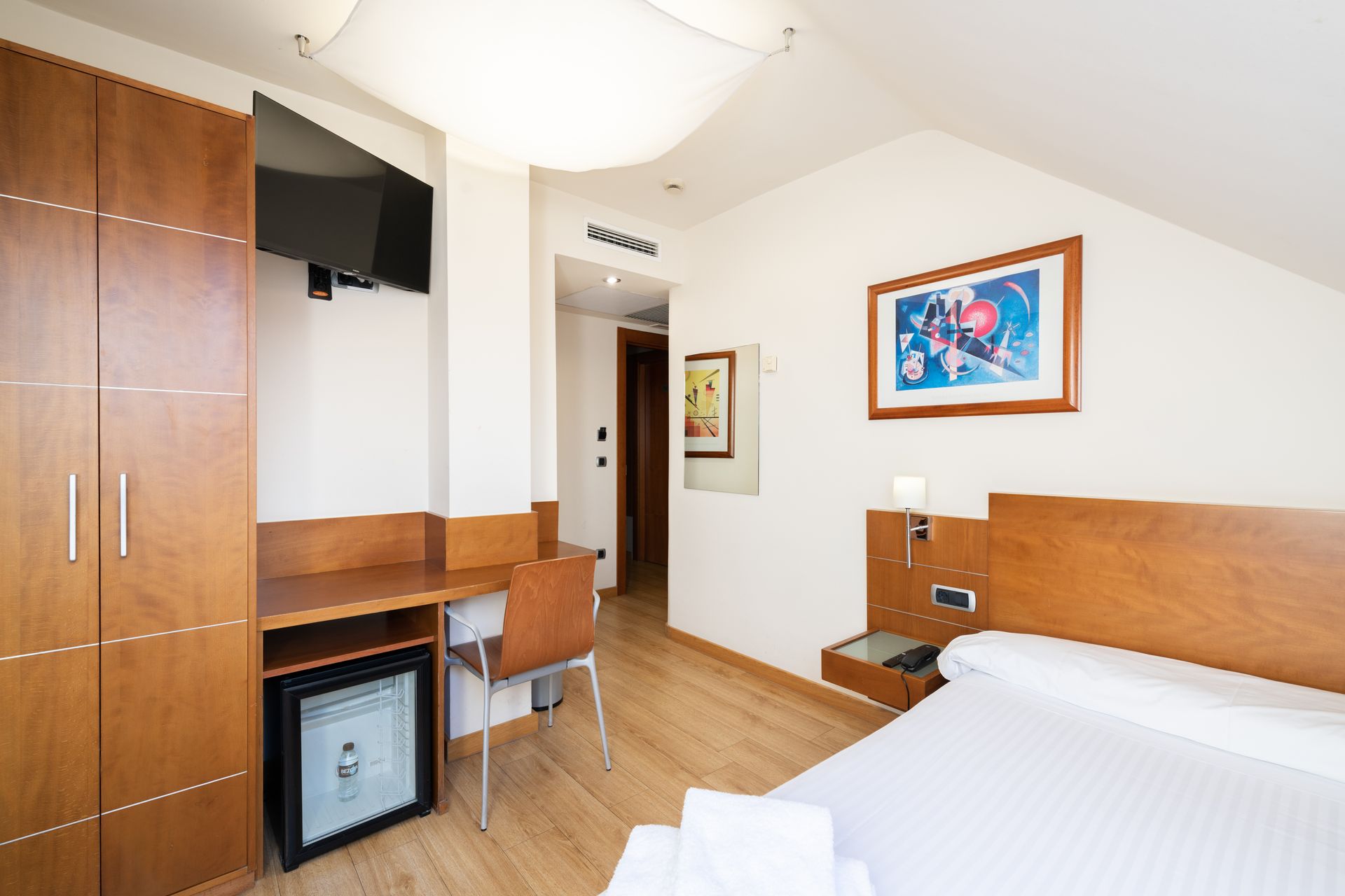 Habitación de hotel luminosa con cama, escritorio de madera, televisor y cuadros enmarcados en la pared.