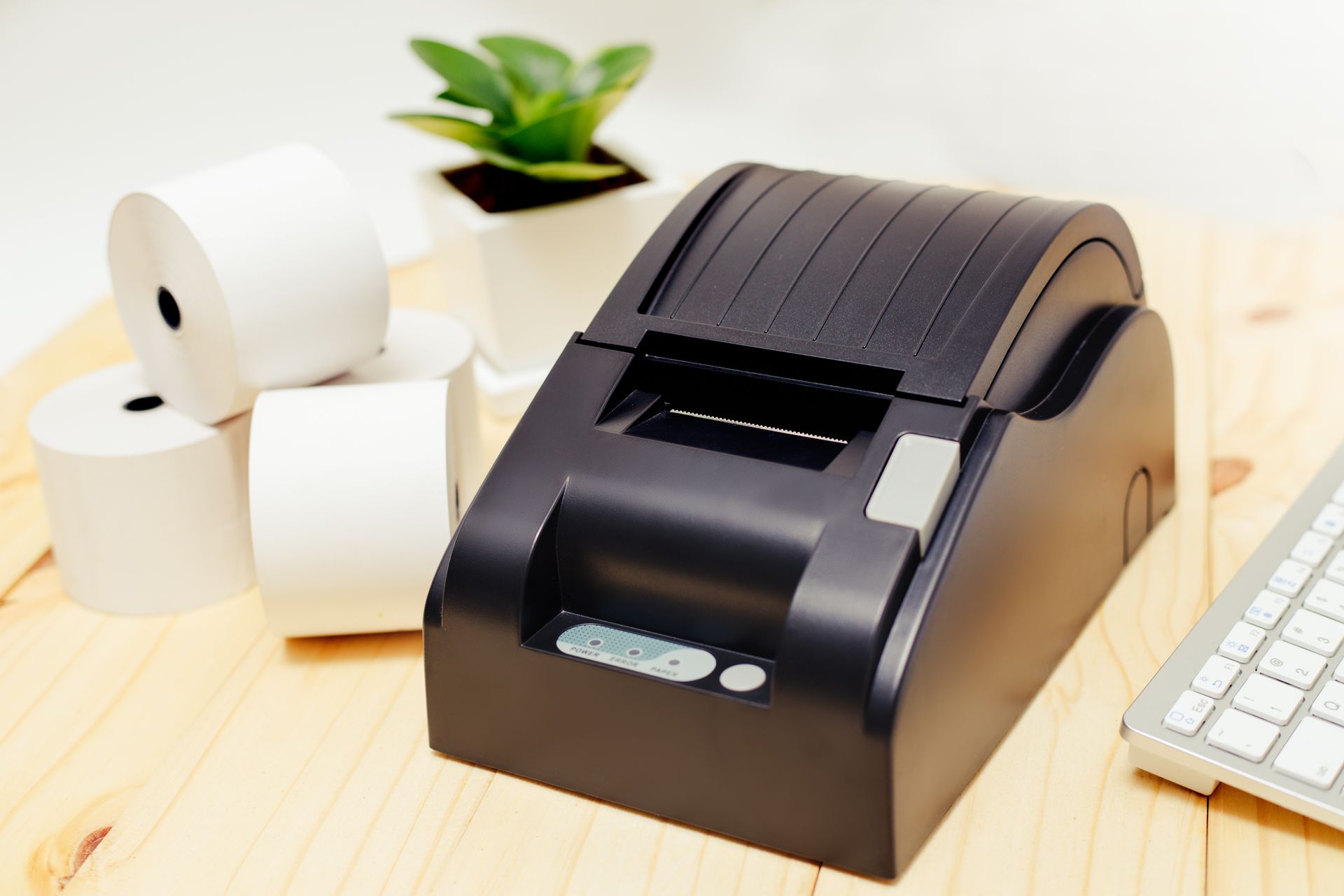 Une imprimante thermique de reçus noire est posée sur un bureau en bois clair, à côté de rouleaux de papier, d'une petite plante en pot et d'un clavier.