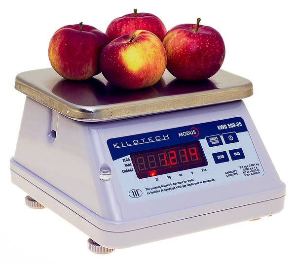 Quatre pommes rouges sont posées sur une balance numérique blanche Kilotech à plateau métallique.