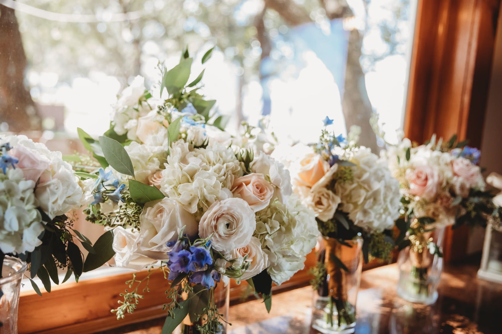 Vases of white and pastel flowers sit on a wood windowsill, with a blurred outdoor backdrop.