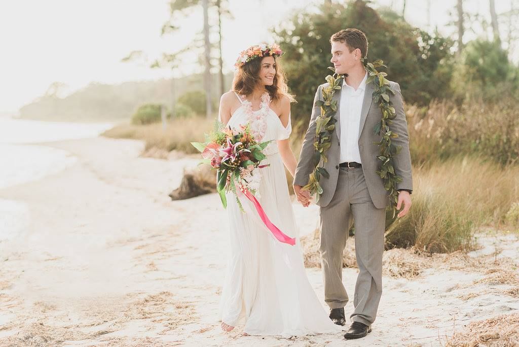 Bride and groom walk hand-in-hand on a beach. She wears a floral crown and holds a bouquet, he wears a greenery lei.