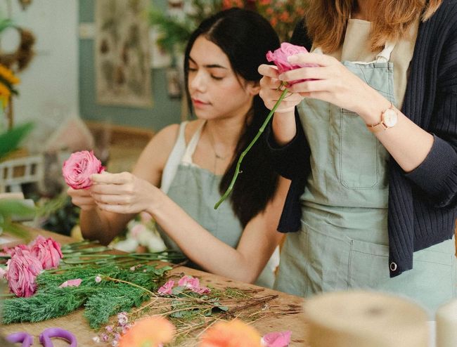 Two people arranging flowers at a table, wearing aprons. One holds a pink rose.