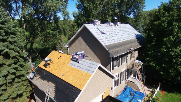 Roofers working on a two-story house, partially covered with new shingles; trees in the background.